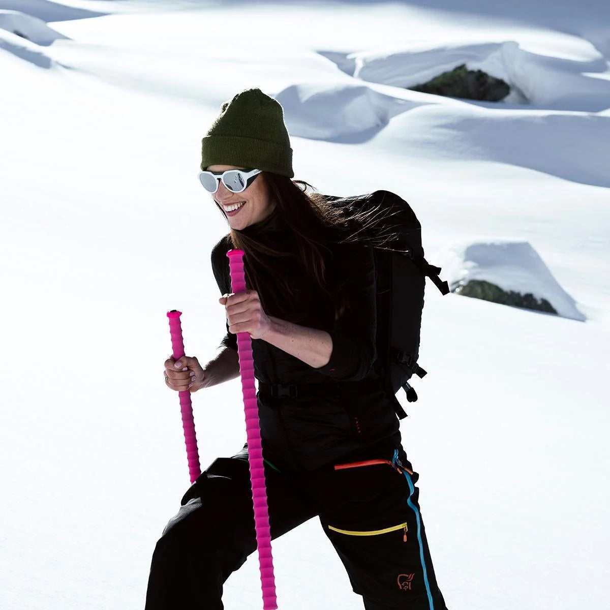 A smiling woman hikes snowy mountains with pink poles, wearing Bolle ASCENDER Sunglasses (Size 51) with removable side shields, a green beanie, and black gear. A smiling woman hikes snowy mountains with pink poles, wearing Bolle ASCENDER Sunglasses (Size 51) with removable side shields, a green beanie, and black gear.