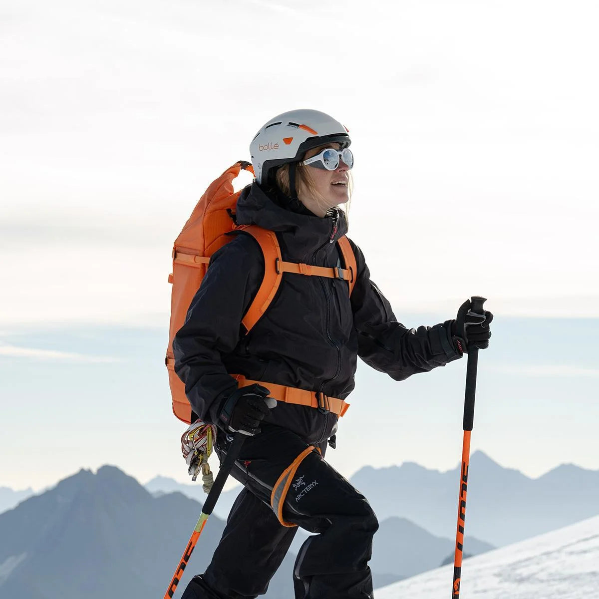 A woman in black ASCENDER gear and helmet hikes snowy mountains with ski poles, wearing Bolle ASCENDER Sunglasses | Size 51 featuring removable side shields and a photochromic lens. Majestic peaks rise in the background. A woman in black ASCENDER gear and helmet hikes snowy mountains with ski poles, wearing Bolle ASCENDER Sunglasses | Size 51 featuring removable side shields and a photochromic lens. Majestic peaks rise in the background.