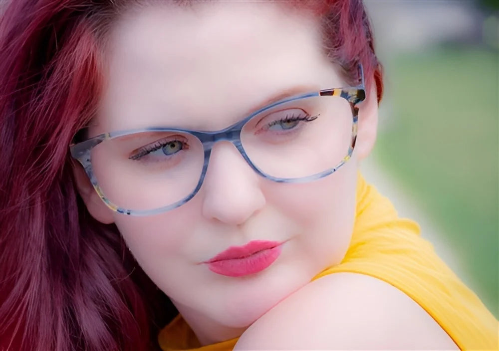 A person with red hair wearing DEA Preferred BARI Eyeglasses (Size 60) and a yellow top is wistfully gazing to the side in a soft-focus outdoor setting. A person with red hair wearing DEA Preferred BARI Eyeglasses (Size 60) and a yellow top is wistfully gazing to the side in a soft-focus outdoor setting.