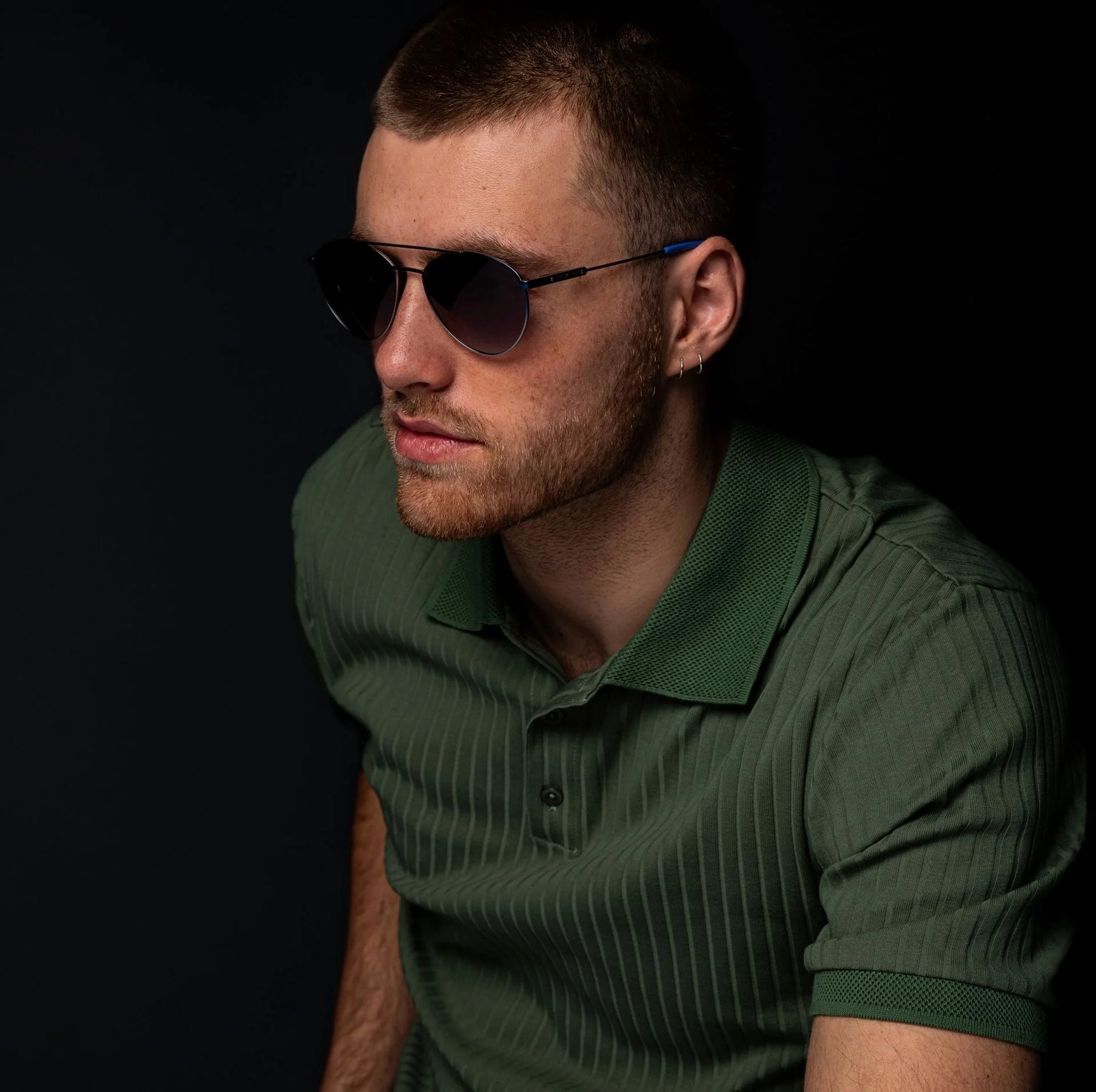 Man in a green polo shirt wearing Di Valdi DV0145 Sunglasses with a geometric shape, seated against a dark background. Man in a green polo shirt wearing Di Valdi DV0145 Sunglasses with a geometric shape, seated against a dark background.