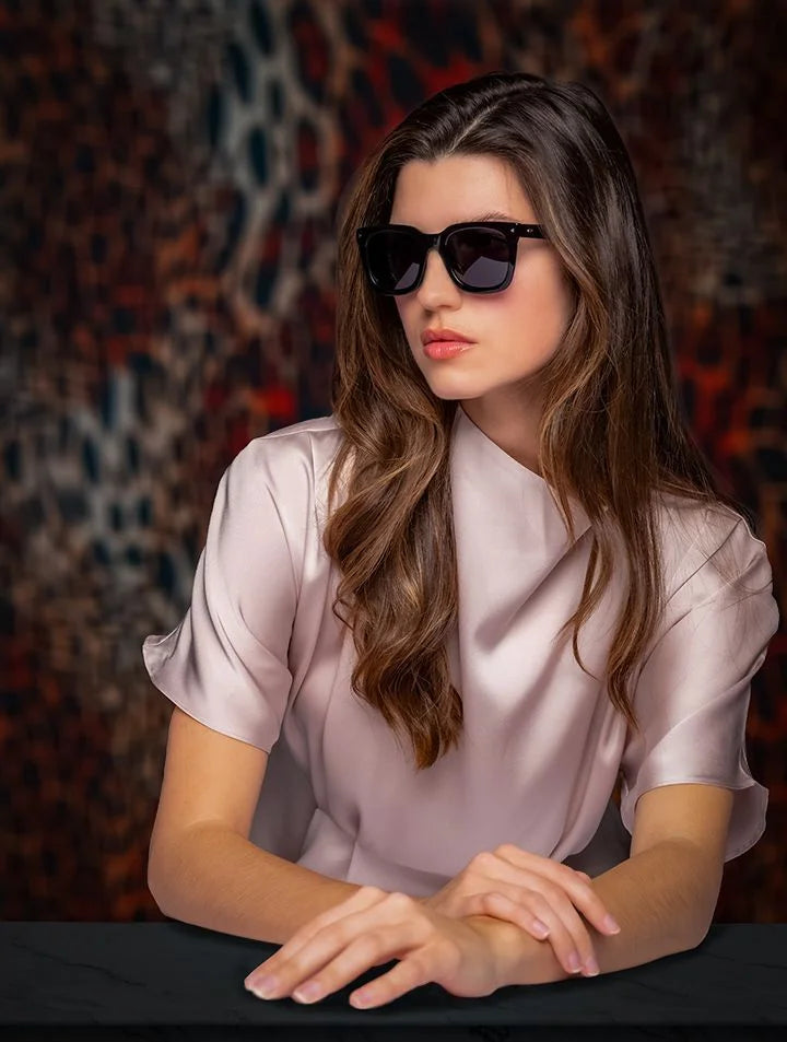 A woman in a light pink blouse wears Di Valdi DV0147 Sunglasses (Size 49) as she sits at a table with her hands folded. A woman in a light pink blouse wears Di Valdi DV0147 Sunglasses (Size 49) as she sits at a table with her hands folded.