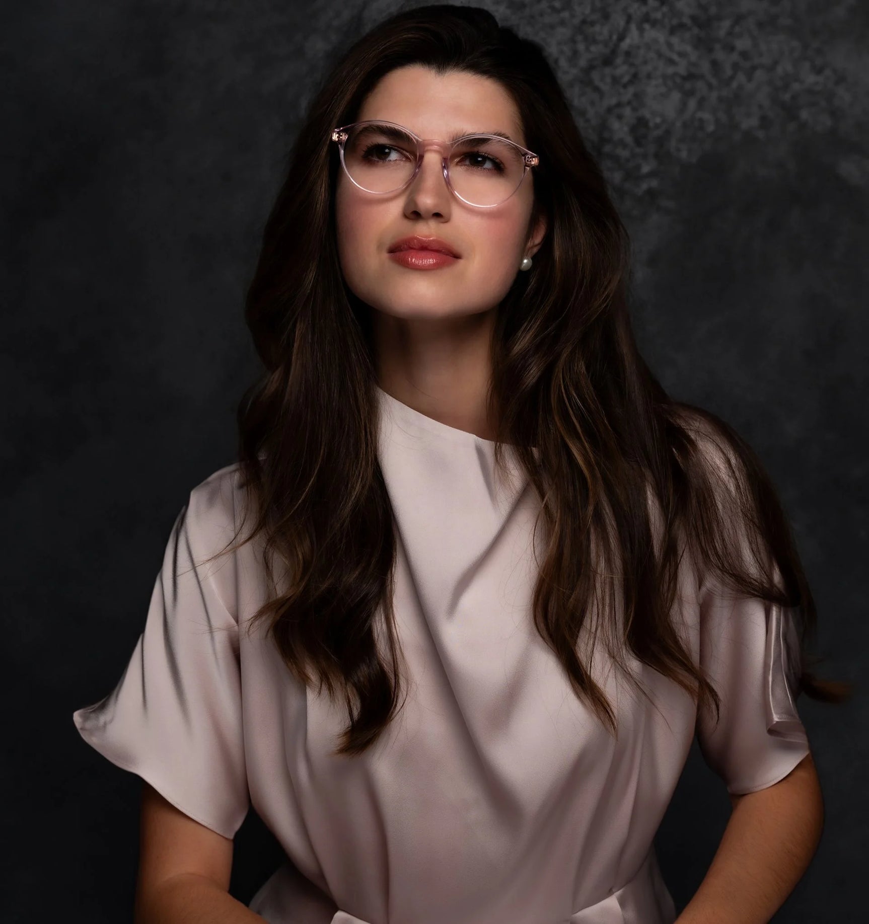 A woman with long brown hair and glasses in a satin blouse looks upward against a dark background, showcasing stylish Di Valdi DVO8181 Eyeglasses | Size 48 by Di Valdi. A woman with long brown hair and glasses in a satin blouse looks upward against a dark background, showcasing stylish Di Valdi DVO8181 Eyeglasses | Size 48 by Di Valdi.