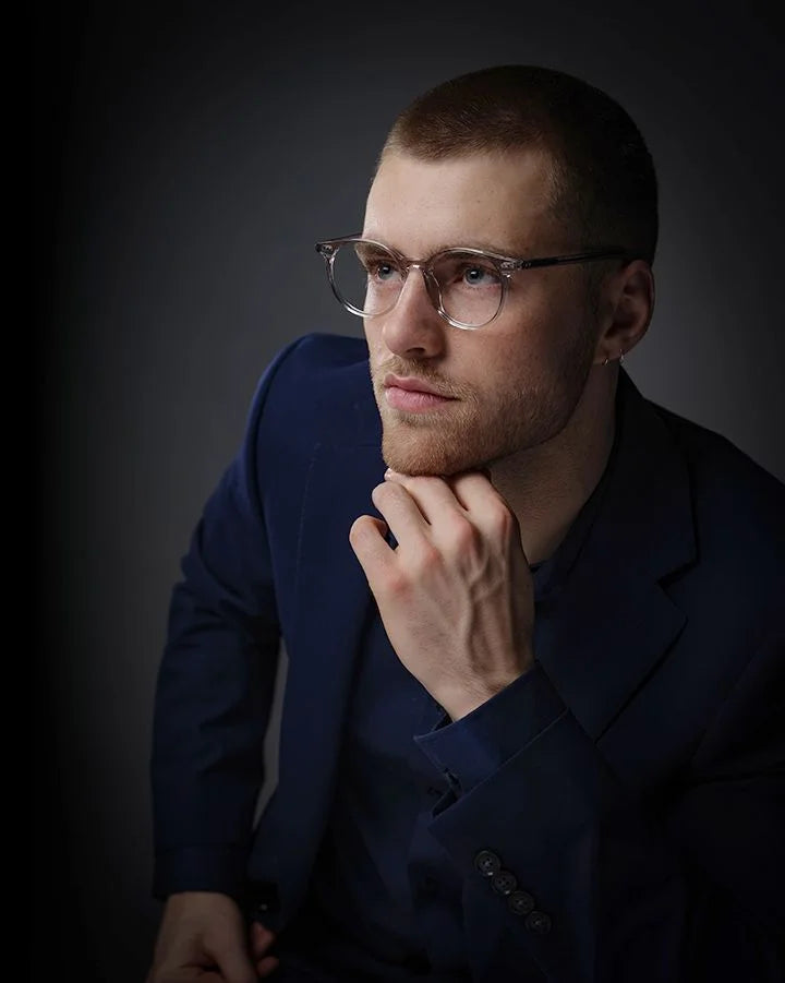 A man in glasses and a dark suit thoughtfully poses with his hand on his chin, highlighting the sleek Di Valdi DVO8186 Eyeglasses | Size 51 by Di Valdi against a dark background. A man in glasses and a dark suit thoughtfully poses with his hand on his chin, highlighting the sleek Di Valdi DVO8186 Eyeglasses | Size 51 by Di Valdi against a dark background.