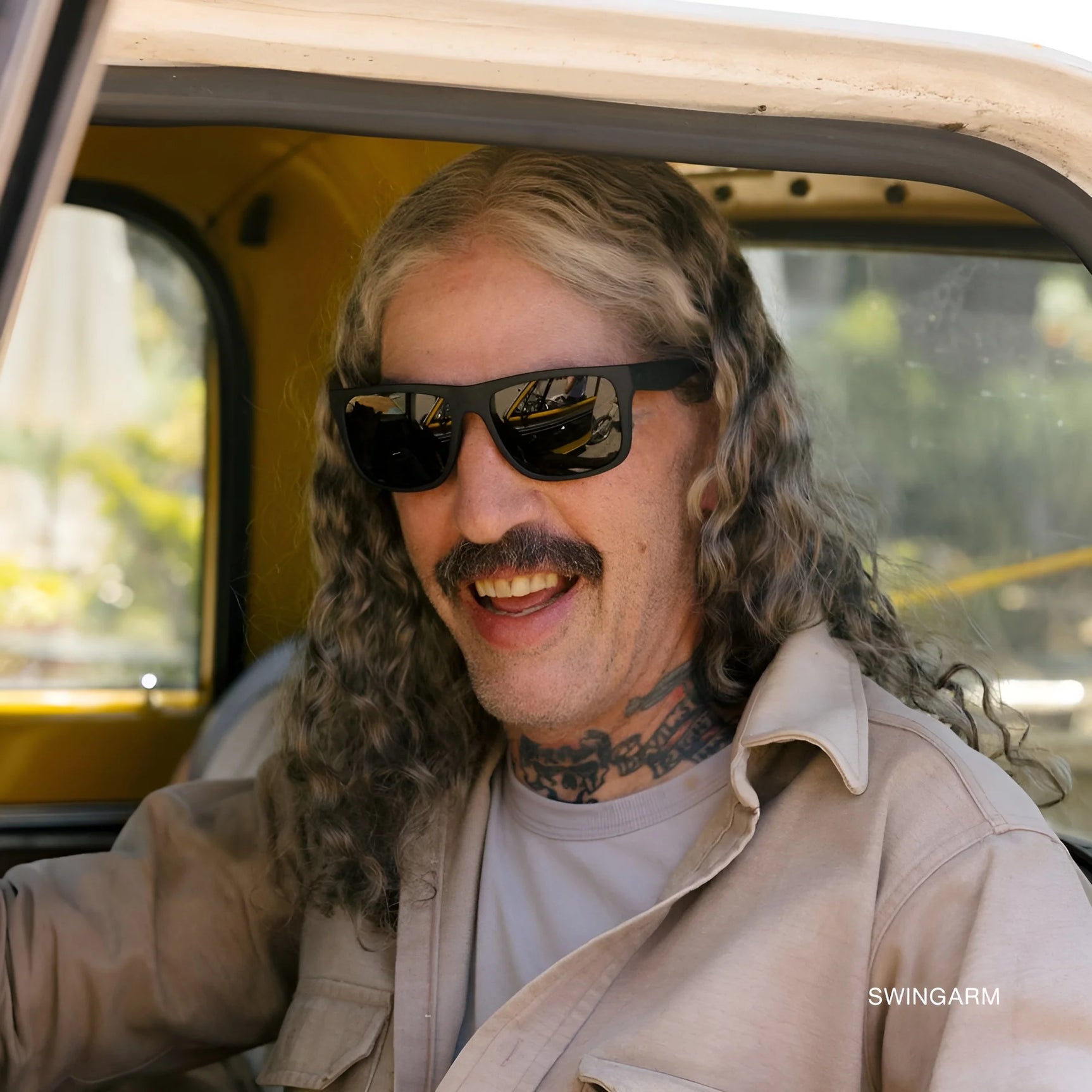 A person with long curly hair wearing Electric Jason Momoa Swingarm M Sunglasses (Size 57) for UV protection smiles while sitting in a vintage yellow car. A person with long curly hair wearing Electric Jason Momoa Swingarm M Sunglasses (Size 57) for UV protection smiles while sitting in a vintage yellow car.
