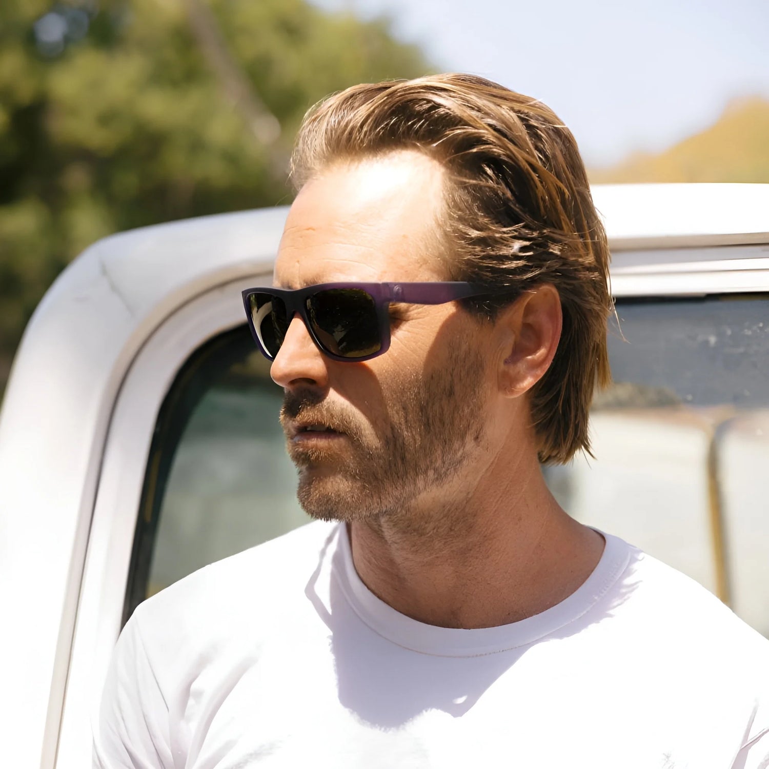 A man wearing Electric Jason Momoa Swingarm M sunglasses and a white shirt stands outside near a vehicle, with trees in the background. A man wearing Electric Jason Momoa Swingarm M sunglasses and a white shirt stands outside near a vehicle, with trees in the background.