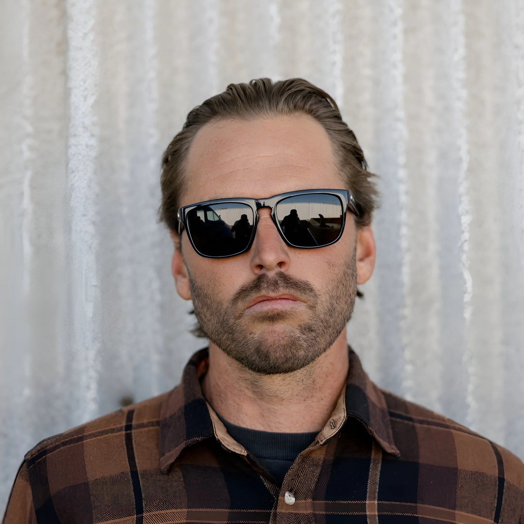 A man in a plaid shirt stands before a corrugated metal wall, sporting Electric Knoxville XL Sunglasses | Size 61 by Electric, highlighting their superior sun protection and lightweight comfort. A man in a plaid shirt stands before a corrugated metal wall, sporting Electric Knoxville XL Sunglasses | Size 61 by Electric, highlighting their superior sun protection and lightweight comfort.