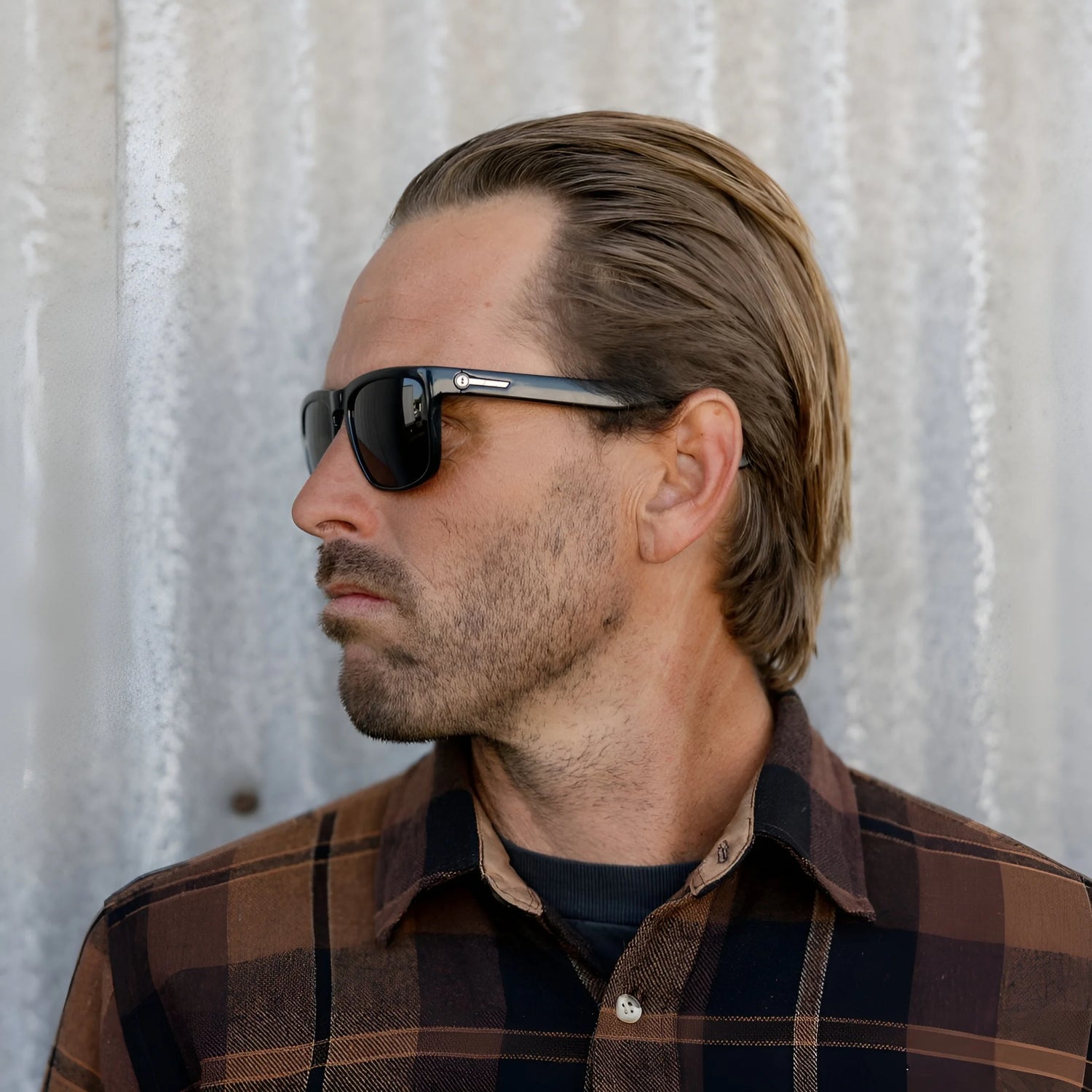 Man with slicked-back hair and Electric Knoxville XL Sunglasses | Size 61 by Electric wears a brown plaid lightweight shirt, facing left against a corrugated wall, highlighting stylish sun protection. Man with slicked-back hair and Electric Knoxville XL Sunglasses | Size 61 by Electric wears a brown plaid lightweight shirt, facing left against a corrugated wall, highlighting stylish sun protection.