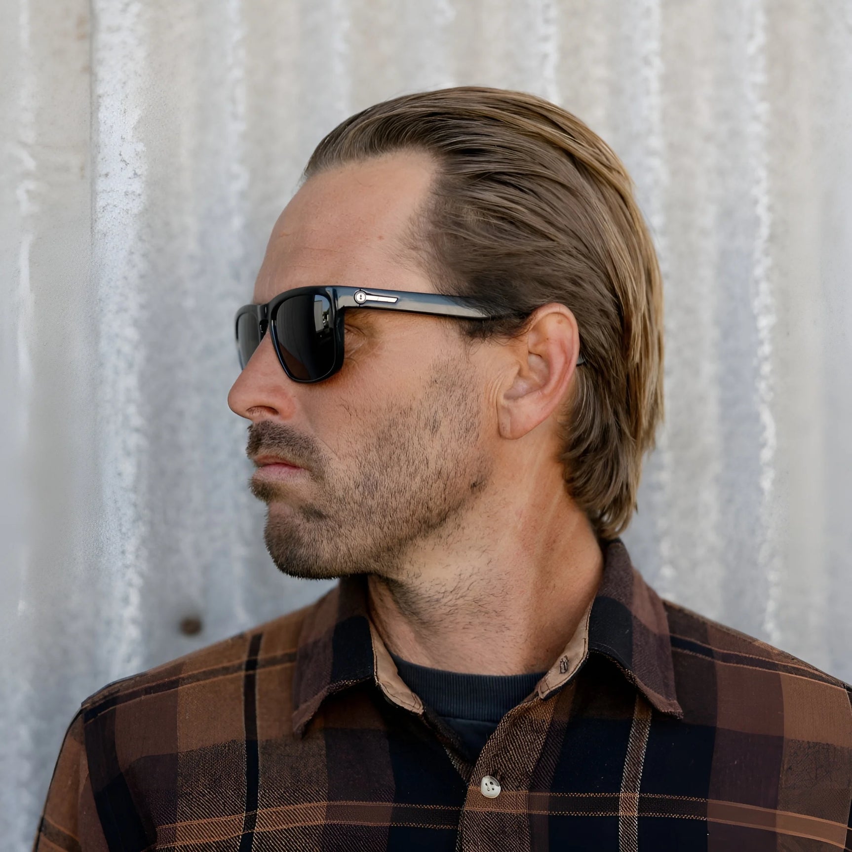 Man with slicked-back hair and Electric Knoxville XL Sunglasses | Size 61 by Electric wears a brown plaid lightweight shirt, facing left against a corrugated wall, highlighting stylish sun protection. Man with slicked-back hair and Electric Knoxville XL Sunglasses | Size 61 by Electric wears a brown plaid lightweight shirt, facing left against a corrugated wall, highlighting stylish sun protection.