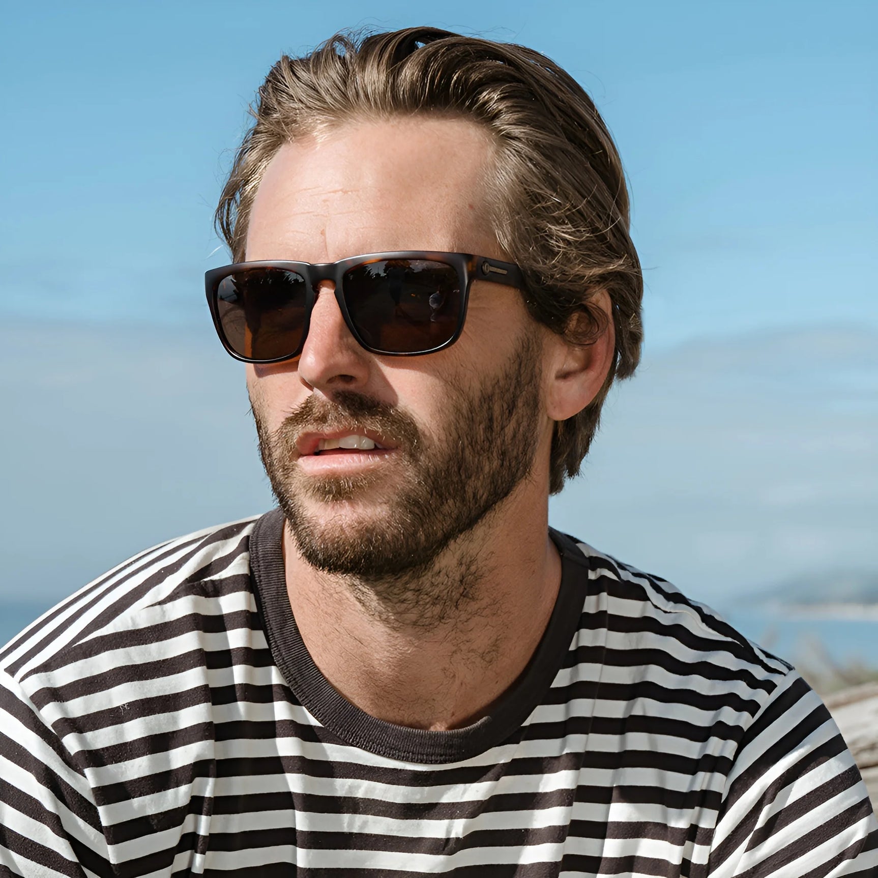 Man with brown hair and beard wears Electric Knoxville XL Sunglasses | Size 61 by Electric and a striped shirt outdoors, enjoying a lightweight fit and excellent sun protection on a sunny day. Man with brown hair and beard wears Electric Knoxville XL Sunglasses | Size 61 by Electric and a striped shirt outdoors, enjoying a lightweight fit and excellent sun protection on a sunny day.
