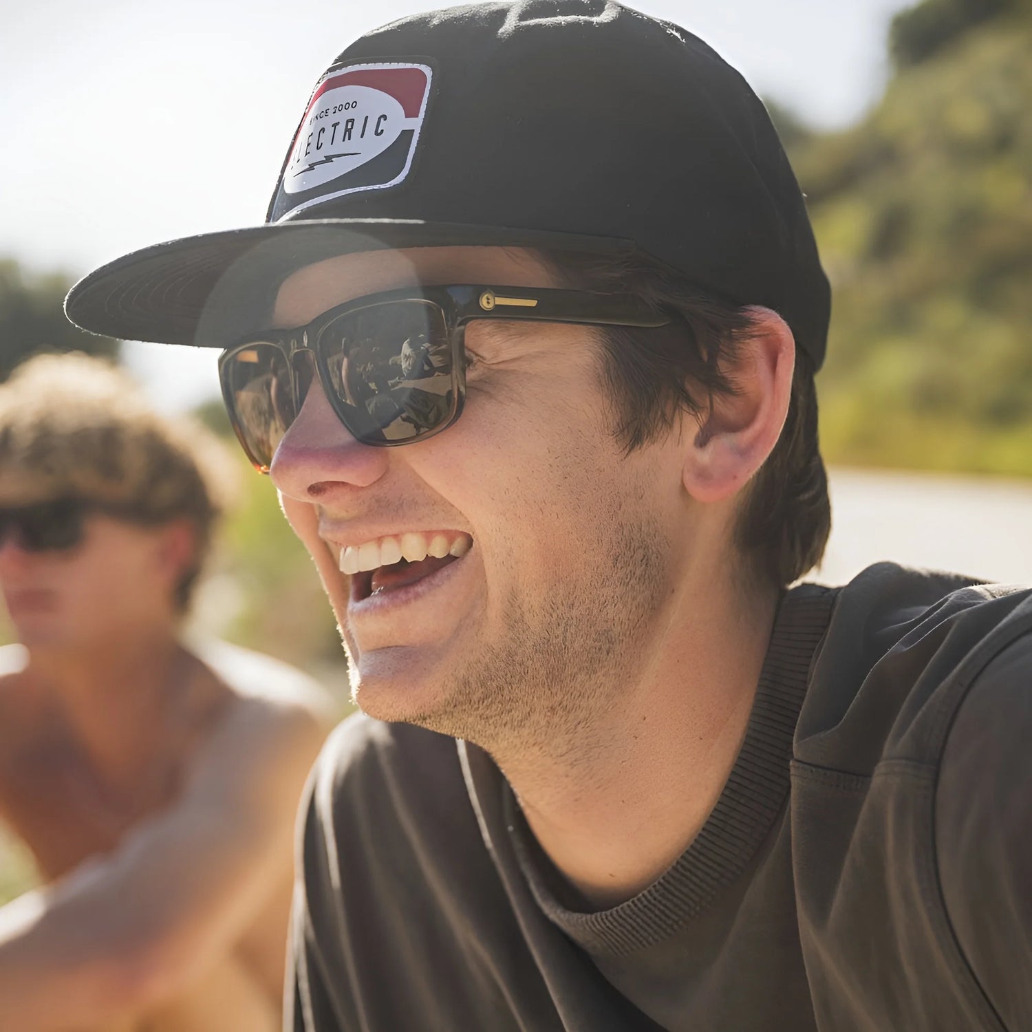 Man wearing Electric Knoxville XL Sunglasses | Size 61 by Electric and a black cap smiles outdoors, enjoying superior sun protection; another person is blurred in the background. Man wearing Electric Knoxville XL Sunglasses | Size 61 by Electric and a black cap smiles outdoors, enjoying superior sun protection; another person is blurred in the background.