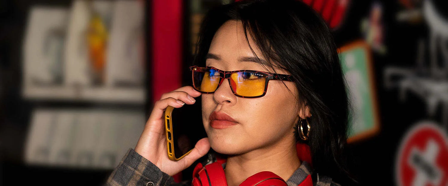 A person wearing Gunnar Cruz Spider-Man Miles Morales Edition Computer Glasses holds a phone to their ear with headphones around their neck, against a blurred background. A person wearing Gunnar Cruz Spider-Man Miles Morales Edition Computer Glasses holds a phone to their ear with headphones around their neck, against a blurred background.