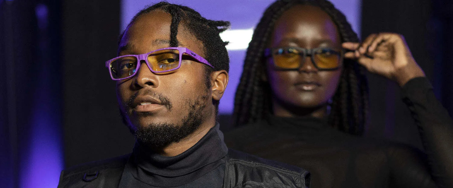 Under purple lighting, two people sport Gunnar's Cruz Black Panther Edition Computer Glasses with durable nylon frames. Their sleek eyewear features Blue Light Protection, perfect for those donning dark attire. One stands in the foreground and one in the background. Under purple lighting, two people sport Gunnar's Cruz Black Panther Edition Computer Glasses with durable nylon frames. Their sleek eyewear features Blue Light Protection, perfect for those donning dark attire. One stands in the foreground and one in the background.