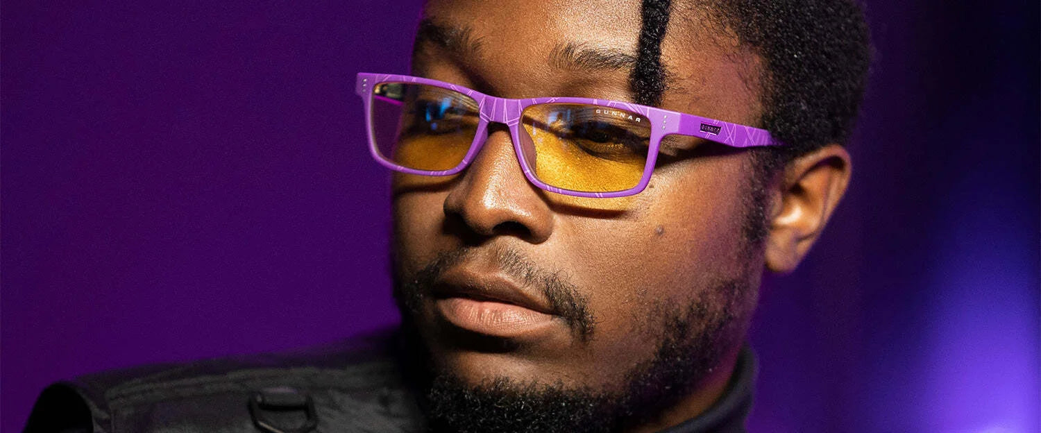 A person with the Gunnar Cruz Black Panther Edition Computer Glasses, featuring a durable nylon frame and yellow lenses for blue light protection, gazes thoughtfully to the side against a purple background. A person with the Gunnar Cruz Black Panther Edition Computer Glasses, featuring a durable nylon frame and yellow lenses for blue light protection, gazes thoughtfully to the side against a purple background.