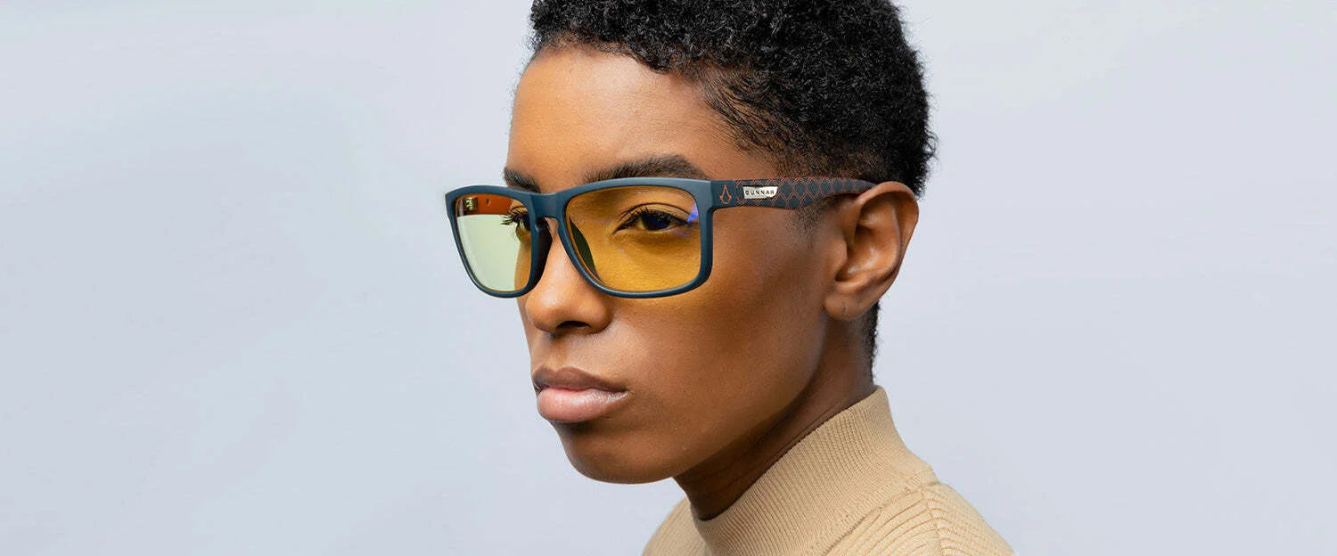 Wearing Gunnar Intercept Assassin’s Creed Mirage Edition Computer Glasses with yellow lenses, a person in a beige sweater stands against a light gray background. These stylish glasses enhance the look while providing excellent blue light protection. Wearing Gunnar Intercept Assassin’s Creed Mirage Edition Computer Glasses with yellow lenses, a person in a beige sweater stands against a light gray background. These stylish glasses enhance the look while providing excellent blue light protection.