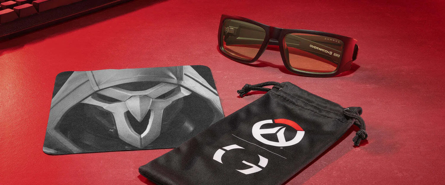Gunnar Overwatch Reaper Computer Glasses with anti-reflective lenses sit stylishly next to a branded pouch and mouse pad on a vibrant red surface. Gunnar Overwatch Reaper Computer Glasses with anti-reflective lenses sit stylishly next to a branded pouch and mouse pad on a vibrant red surface.
