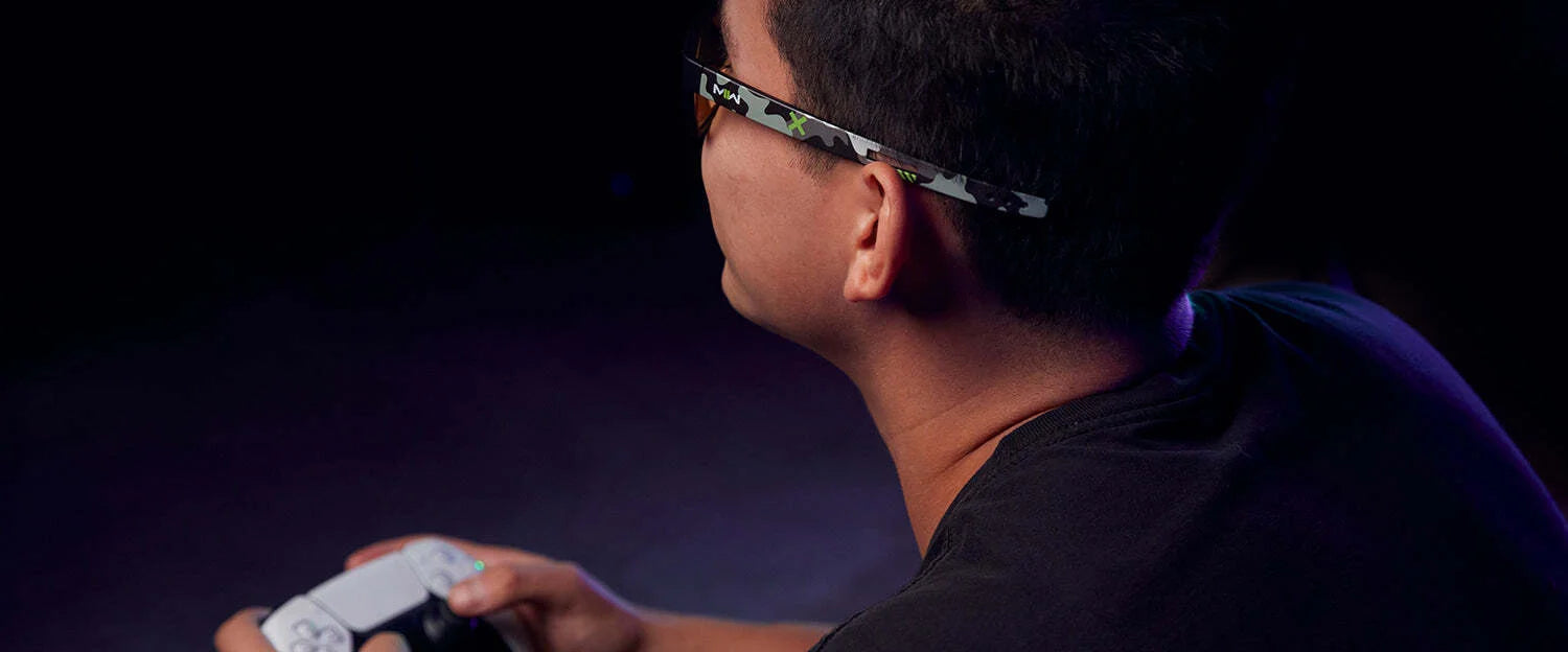 In a dimly lit room, a person wears Gunnar Call of Duty Tactical Edition Computer Glasses, gripping a gaming controller with focus and readiness for action. In a dimly lit room, a person wears Gunnar Call of Duty Tactical Edition Computer Glasses, gripping a gaming controller with focus and readiness for action.