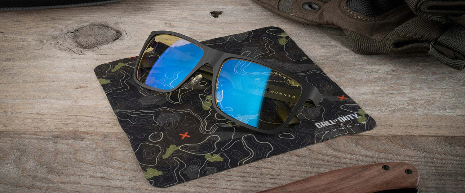 Gunnar Call of Duty UAV Edition Computer Glasses with blue lenses rest on a themed cloth atop a wooden surface. These glasses, by Gunnar, feature durable and stylish aluminum-magnesium forged frames. Gunnar Call of Duty UAV Edition Computer Glasses with blue lenses rest on a themed cloth atop a wooden surface. These glasses, by Gunnar, feature durable and stylish aluminum-magnesium forged frames.