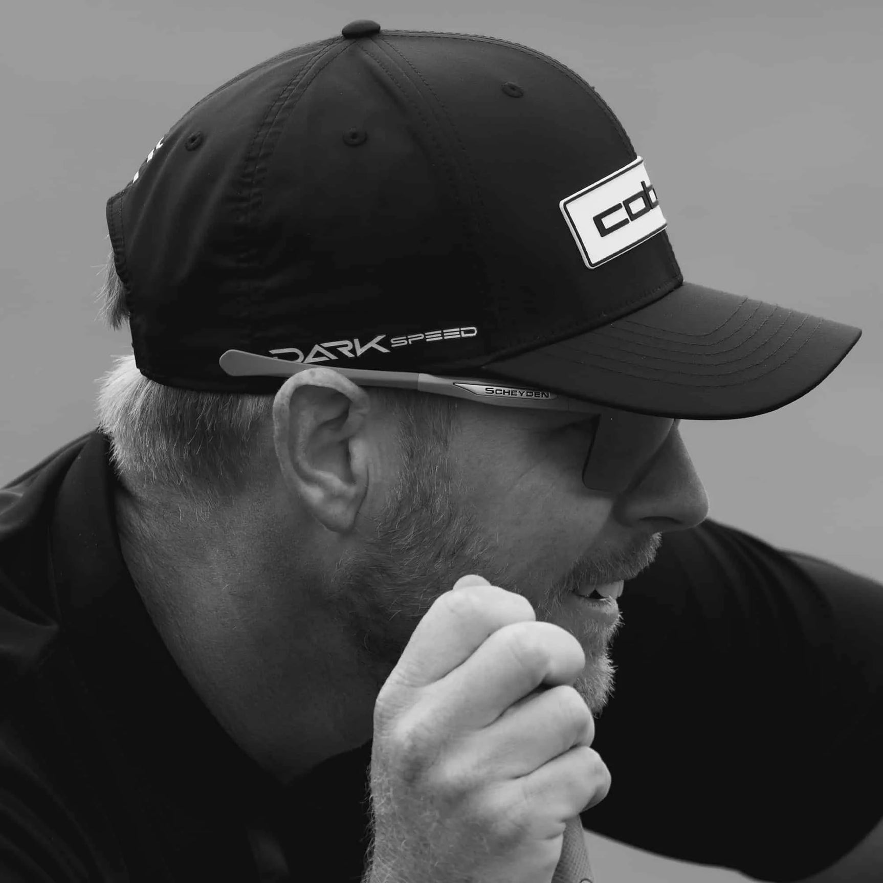 A man in a cap and Scheyden CIA GRABBER SMALL Sunglasses (Size 58) holds a golf club, looking focused. The black-and-white image highlights the stylish eyewear by Scheyden. A man in a cap and Scheyden CIA GRABBER SMALL Sunglasses (Size 58) holds a golf club, looking focused. The black-and-white image highlights the stylish eyewear by Scheyden.