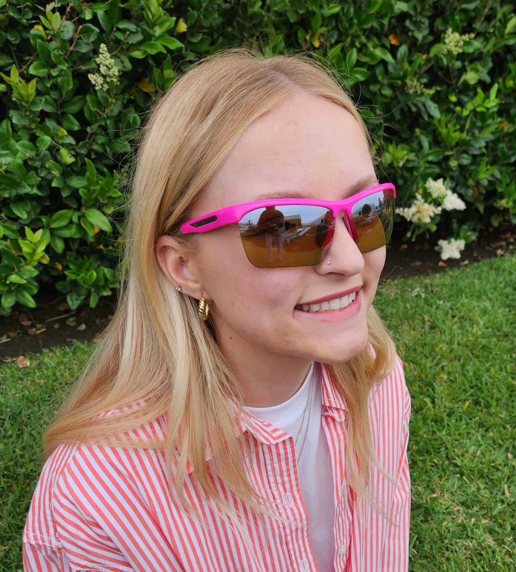 A smiling woman with blonde hair, wearing Scheyden CIA GRABBER SMALL Sunglasses (Size 58) and a striped shirt, sits on grass near green bushes. A smiling woman with blonde hair, wearing Scheyden CIA GRABBER SMALL Sunglasses (Size 58) and a striped shirt, sits on grass near green bushes.