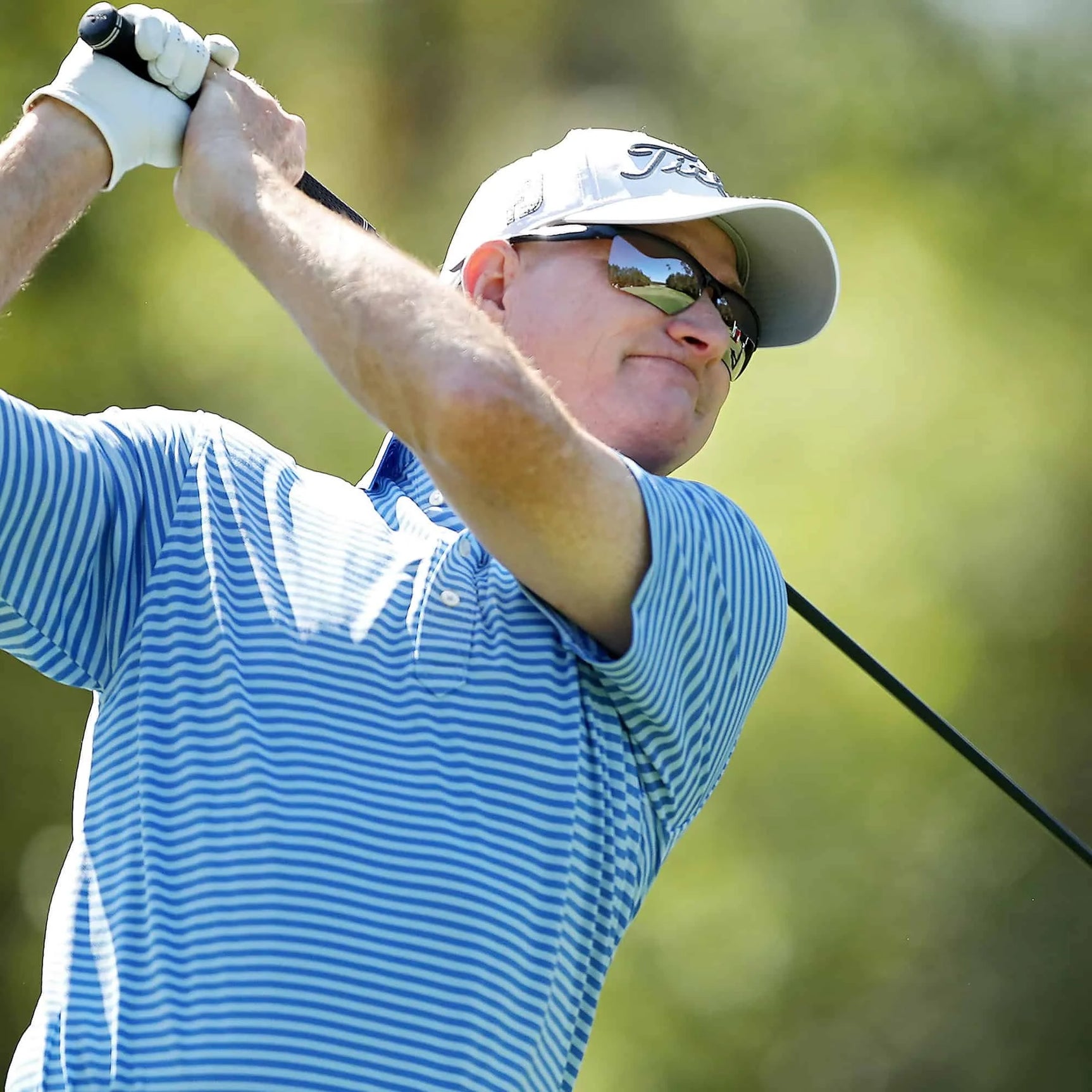 A golfer in a blue striped shirt swings a club outdoors on a sunny day, wearing Scheyden CIA GRABBER SMALL Sunglasses | Size 58 with LT33 RSR lenses and TR90 composite frame for optimal clarity and comfort. A golfer in a blue striped shirt swings a club outdoors on a sunny day, wearing Scheyden CIA GRABBER SMALL Sunglasses | Size 58 with LT33 RSR lenses and TR90 composite frame for optimal clarity and comfort.