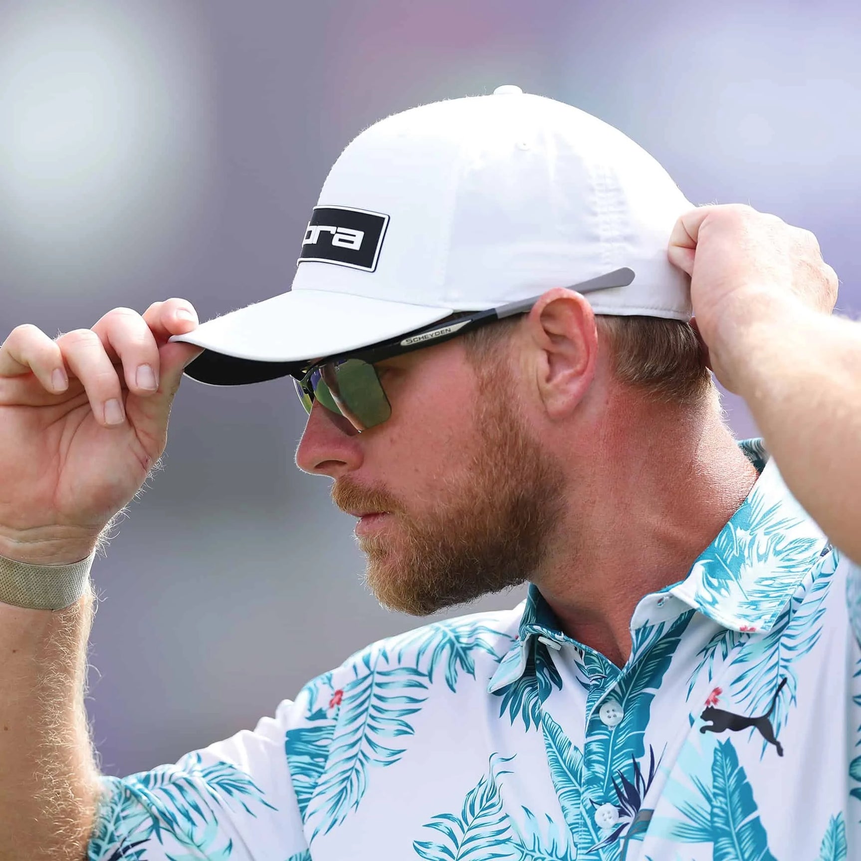 A man in a patterned palm leaf shirt adjusts his white cap while wearing Scheyden CIA GRABBER Sunglasses, size 65. A man in a patterned palm leaf shirt adjusts his white cap while wearing Scheyden CIA GRABBER Sunglasses, size 65.