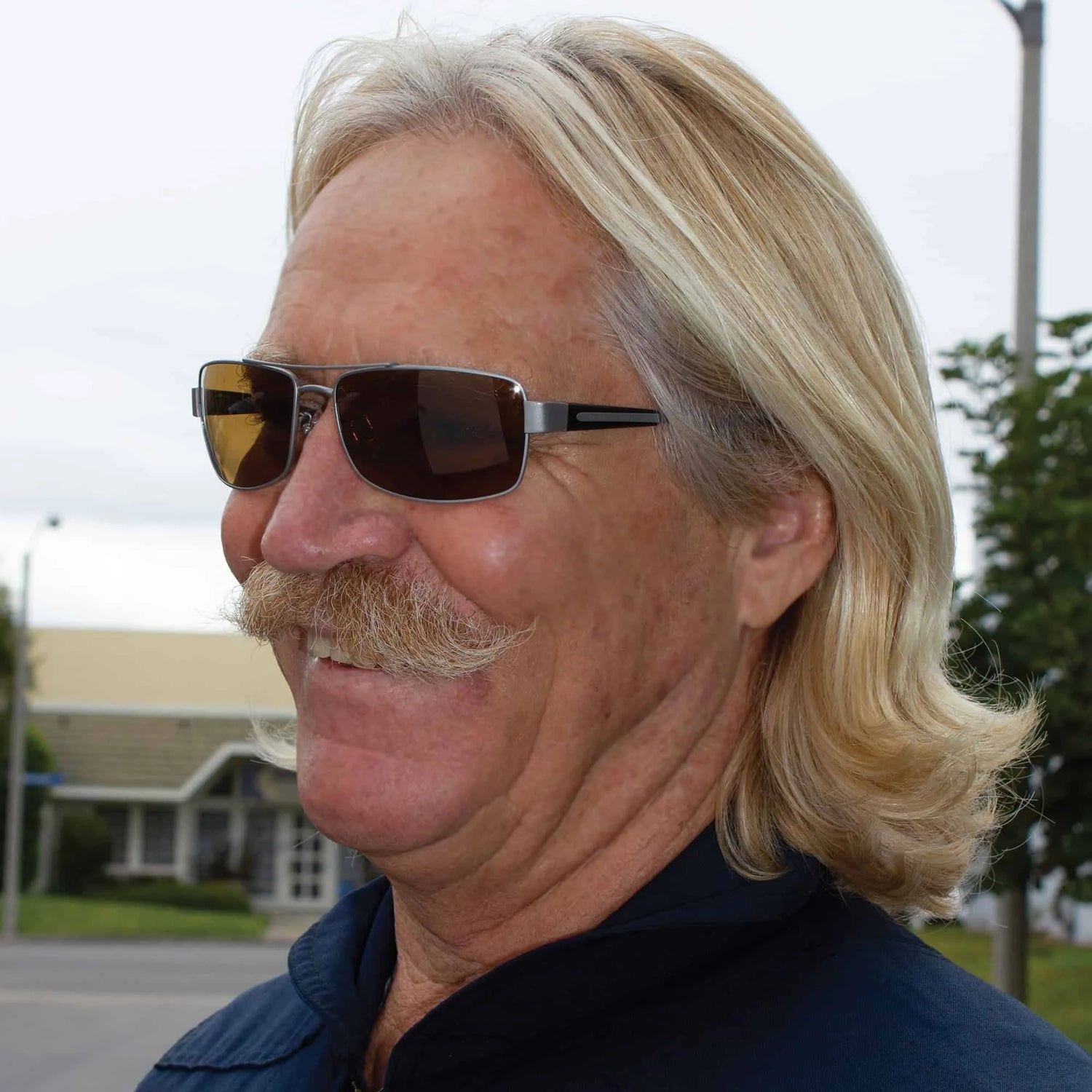 A blonde man with a bushy mustache, wearing Scheyden MUSTANG Sunglasses, Size 60, featuring polarized lenses, is smiling outdoors. A blonde man with a bushy mustache, wearing Scheyden MUSTANG Sunglasses, Size 60, featuring polarized lenses, is smiling outdoors.