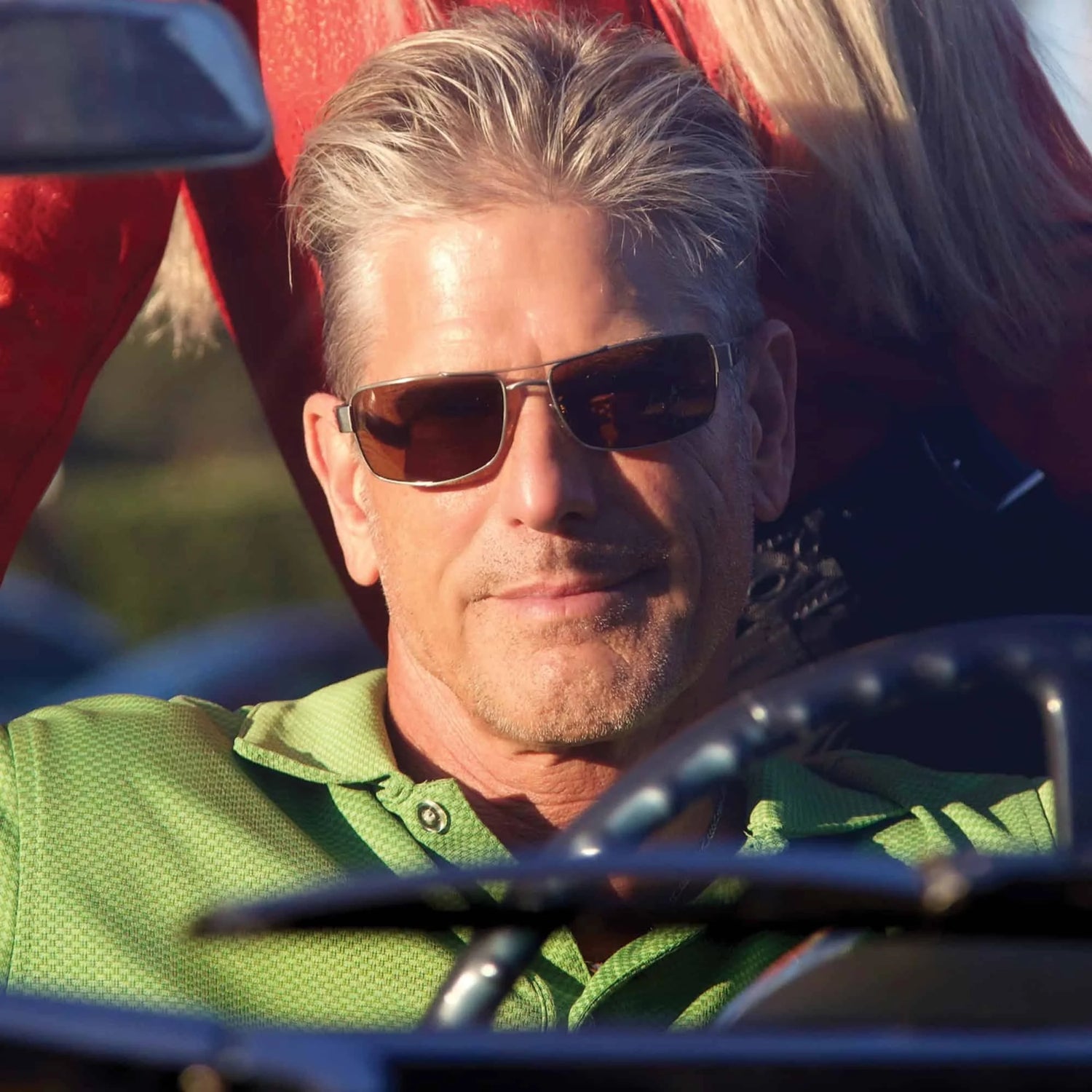 A man wearing Scheyden MUSTANG sunglasses with polarized lenses and a green shirt drives a convertible, smiling slightly under the bright sunlight. A man wearing Scheyden MUSTANG sunglasses with polarized lenses and a green shirt drives a convertible, smiling slightly under the bright sunlight.