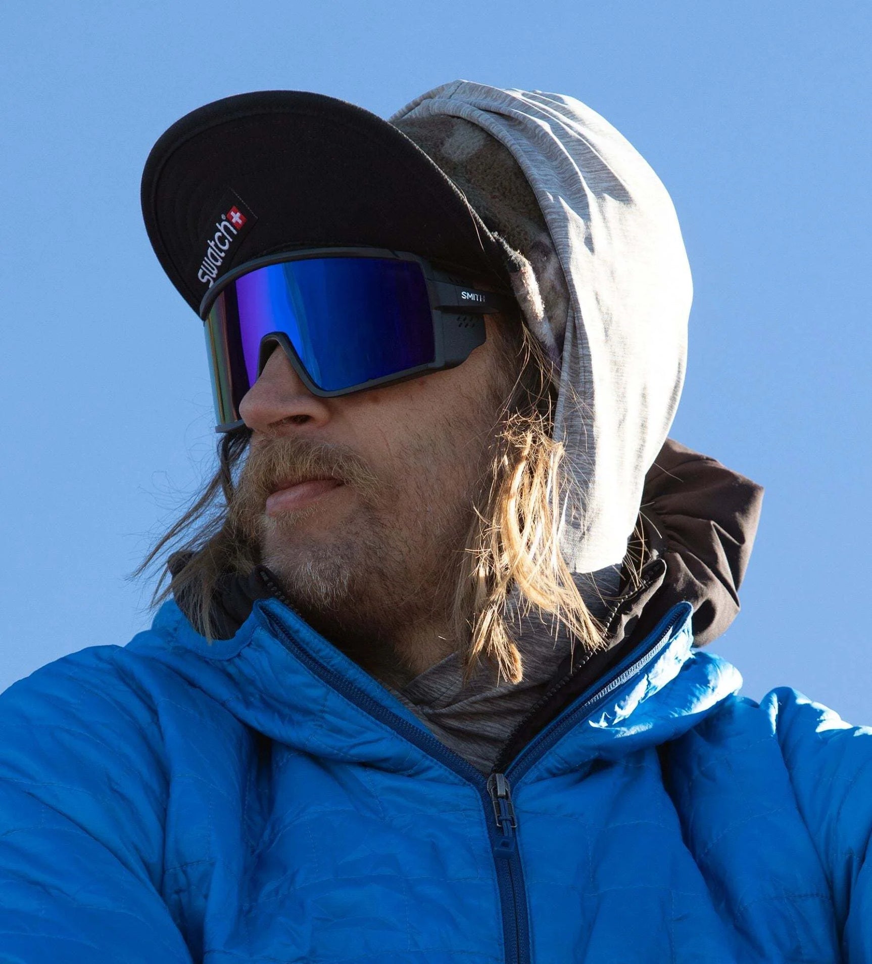A person in a blue jacket, wearing reflective Smith Optics Pursuit Sunglasses along with a cap and hood, gazes to the side against a striking blue sky. A person in a blue jacket, wearing reflective Smith Optics Pursuit Sunglasses along with a cap and hood, gazes to the side against a striking blue sky.