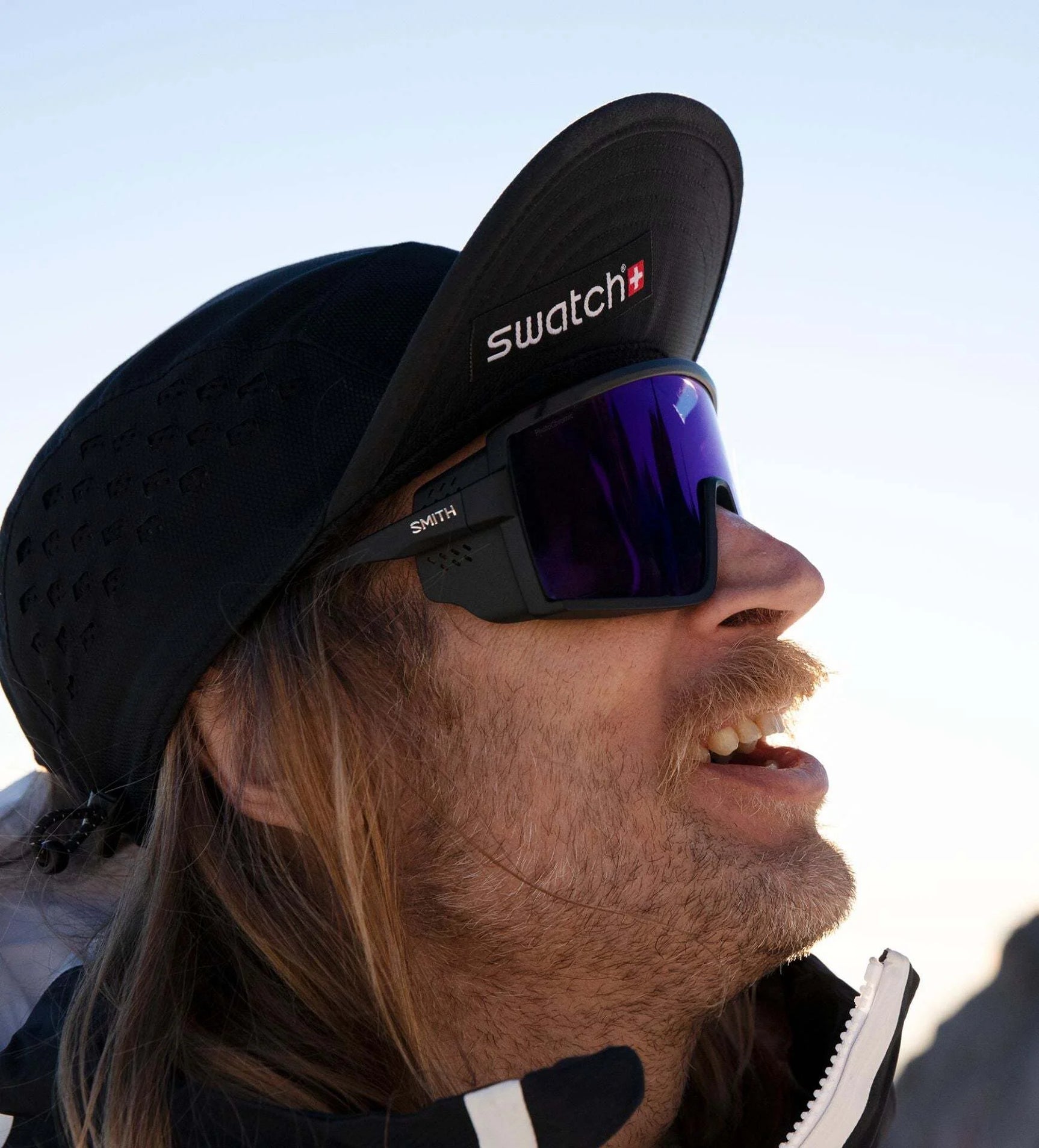 Person wearing a cap and Smith Optics Pursuit Sunglasses, looking upward with a smile against a clear sky backdrop. Person wearing a cap and Smith Optics Pursuit Sunglasses, looking upward with a smile against a clear sky backdrop.