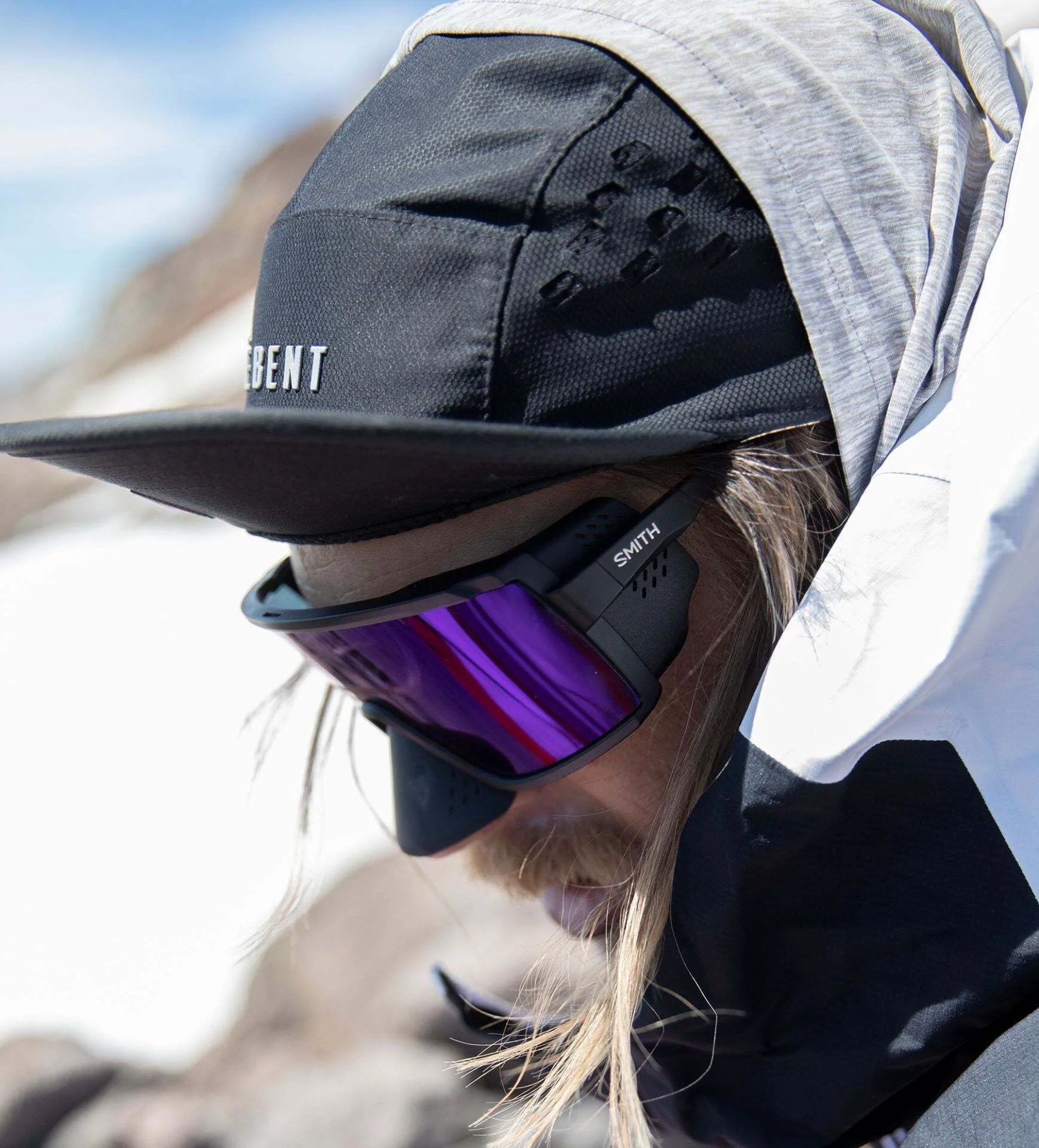 A person in winter clothing equipped with Smith Optics Pursuit Sunglasses, featuring photochromic lenses, stands outdoors under a hat and hood amidst snow-covered terrain. A person in winter clothing equipped with Smith Optics Pursuit Sunglasses, featuring photochromic lenses, stands outdoors under a hat and hood amidst snow-covered terrain.