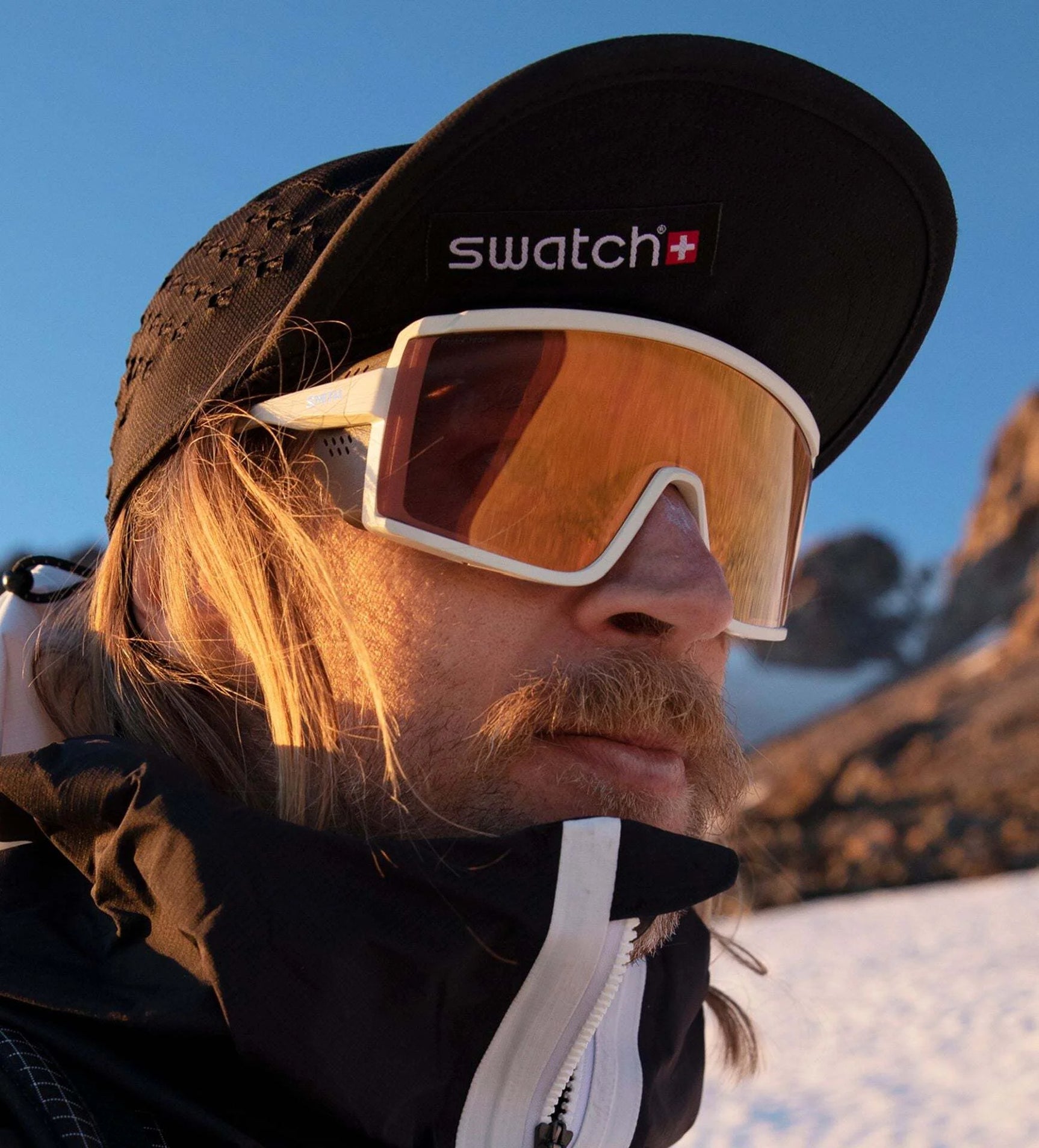 A person wearing a cap and Smith Optics Pursuit Sunglasses with reflective Glacier Photochromic lenses stands against a backdrop of majestic mountains, ready for adventure. A person wearing a cap and Smith Optics Pursuit Sunglasses with reflective Glacier Photochromic lenses stands against a backdrop of majestic mountains, ready for adventure.