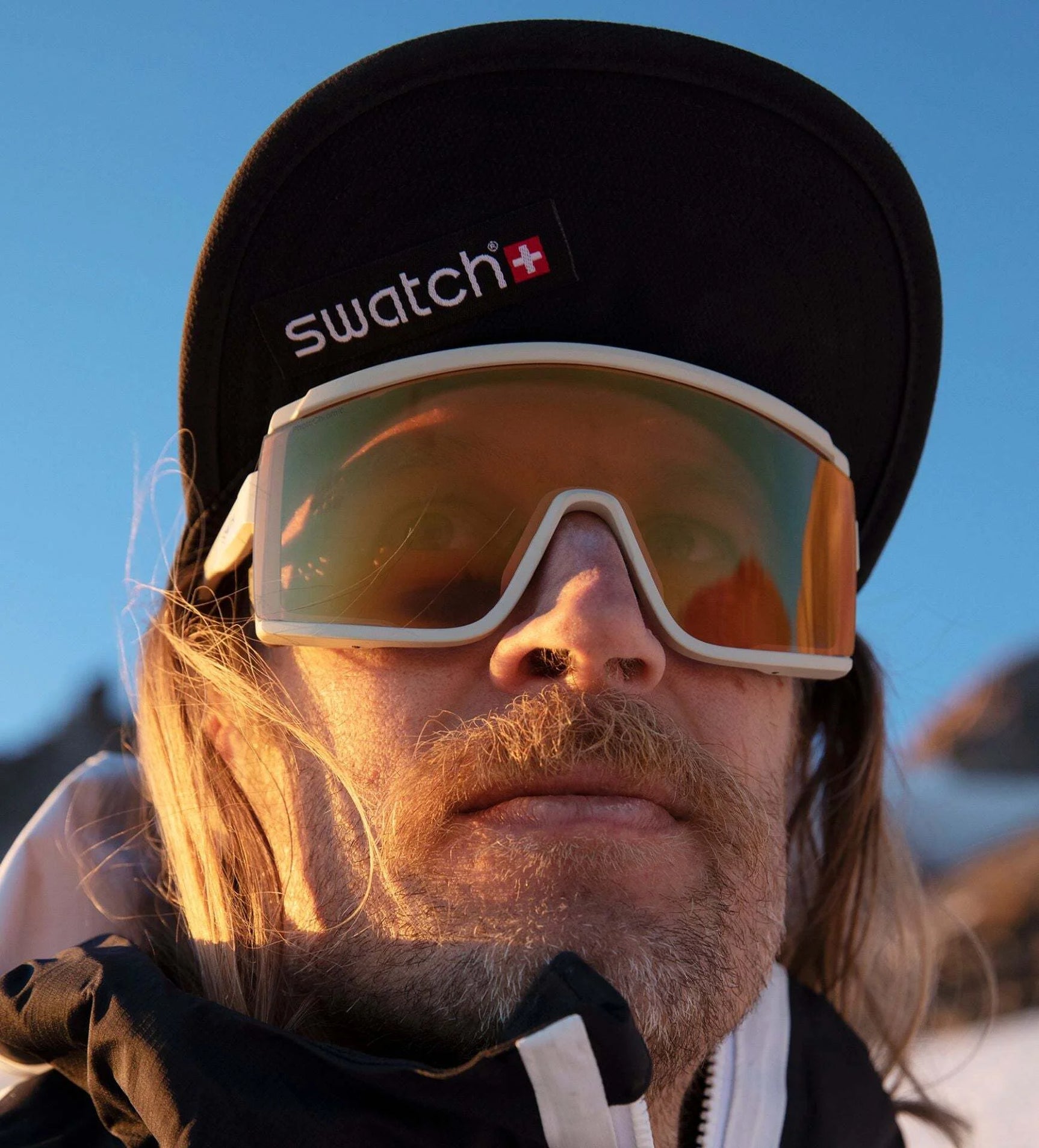 A person with long hair wearing large Smith Optics Pursuit Sunglasses, which feature photochromic lenses, and a Swatch cap poses against a stunning mountainous backdrop. A person with long hair wearing large Smith Optics Pursuit Sunglasses, which feature photochromic lenses, and a Swatch cap poses against a stunning mountainous backdrop.