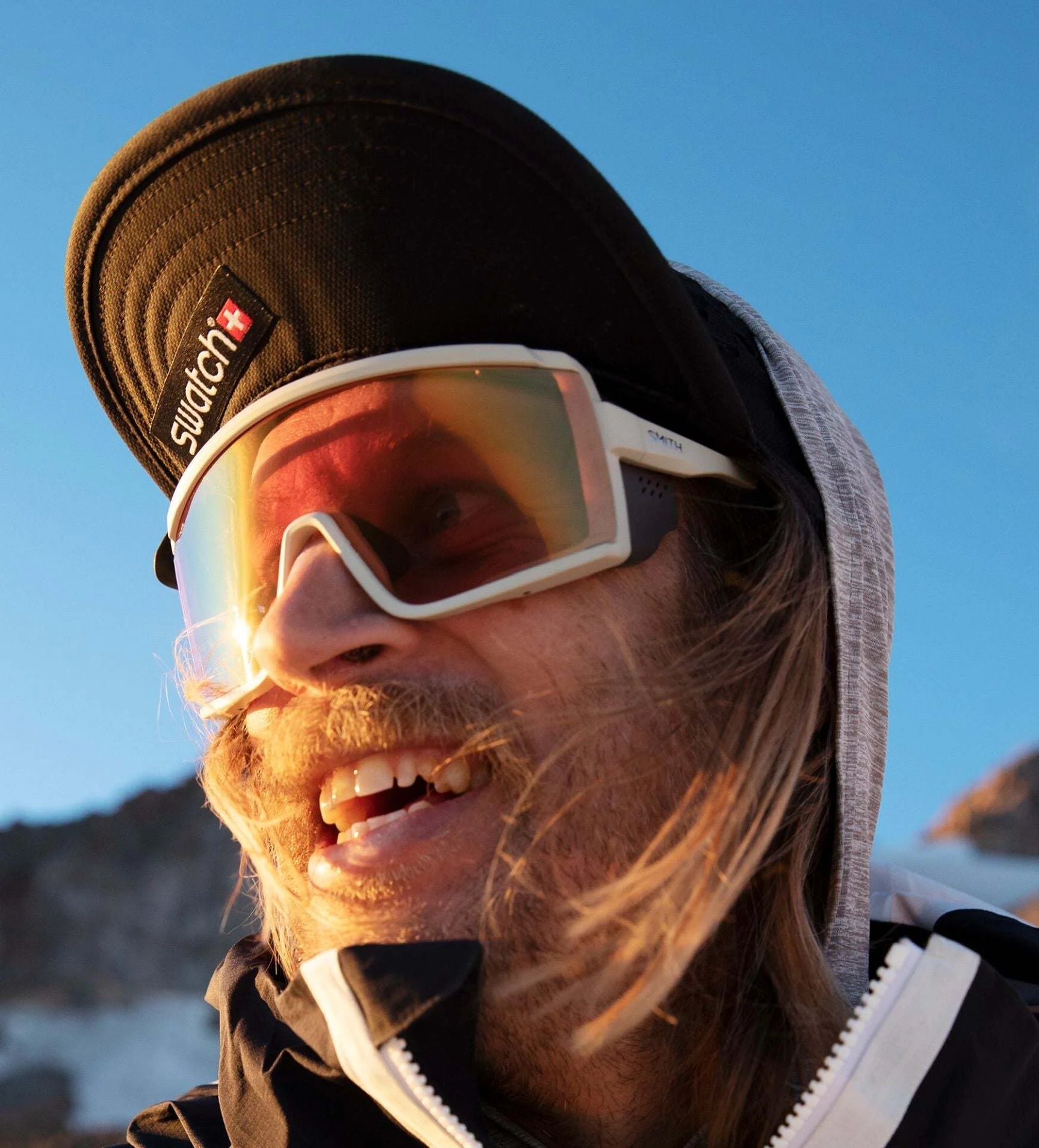 A person with long hair wearing Smith Optics Pursuit Sunglasses, a cap, and a hoodie smiles outdoors under the clear blue sky. A person with long hair wearing Smith Optics Pursuit Sunglasses, a cap, and a hoodie smiles outdoors under the clear blue sky.