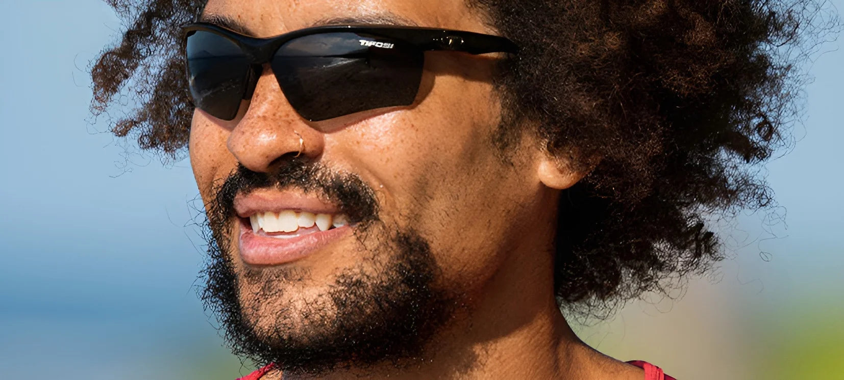 A curly-haired person with a smaller face smiles outdoors, enjoying the sunshine in their Tifosi Optics VERO Sunglasses, Size 64. A curly-haired person with a smaller face smiles outdoors, enjoying the sunshine in their Tifosi Optics VERO Sunglasses, Size 64.