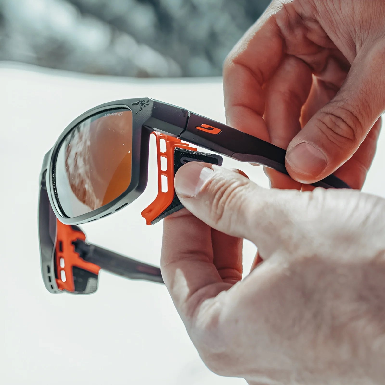 Hands adjusting the side shields on Julbo Shield Sunglasses in black and orange with reflective lenses, featuring Grip Nose technology for a secure fit. Hands adjusting the side shields on Julbo Shield Sunglasses in black and orange with reflective lenses, featuring Grip Nose technology for a secure fit.