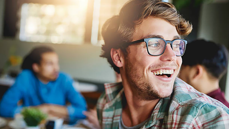 Smiling man with glasses and plaid shirt looks to the side; blurred people in the background.