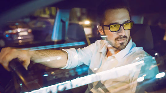 Man wearing yellow-tinted glasses driving at night, city lights reflecting on the car window.