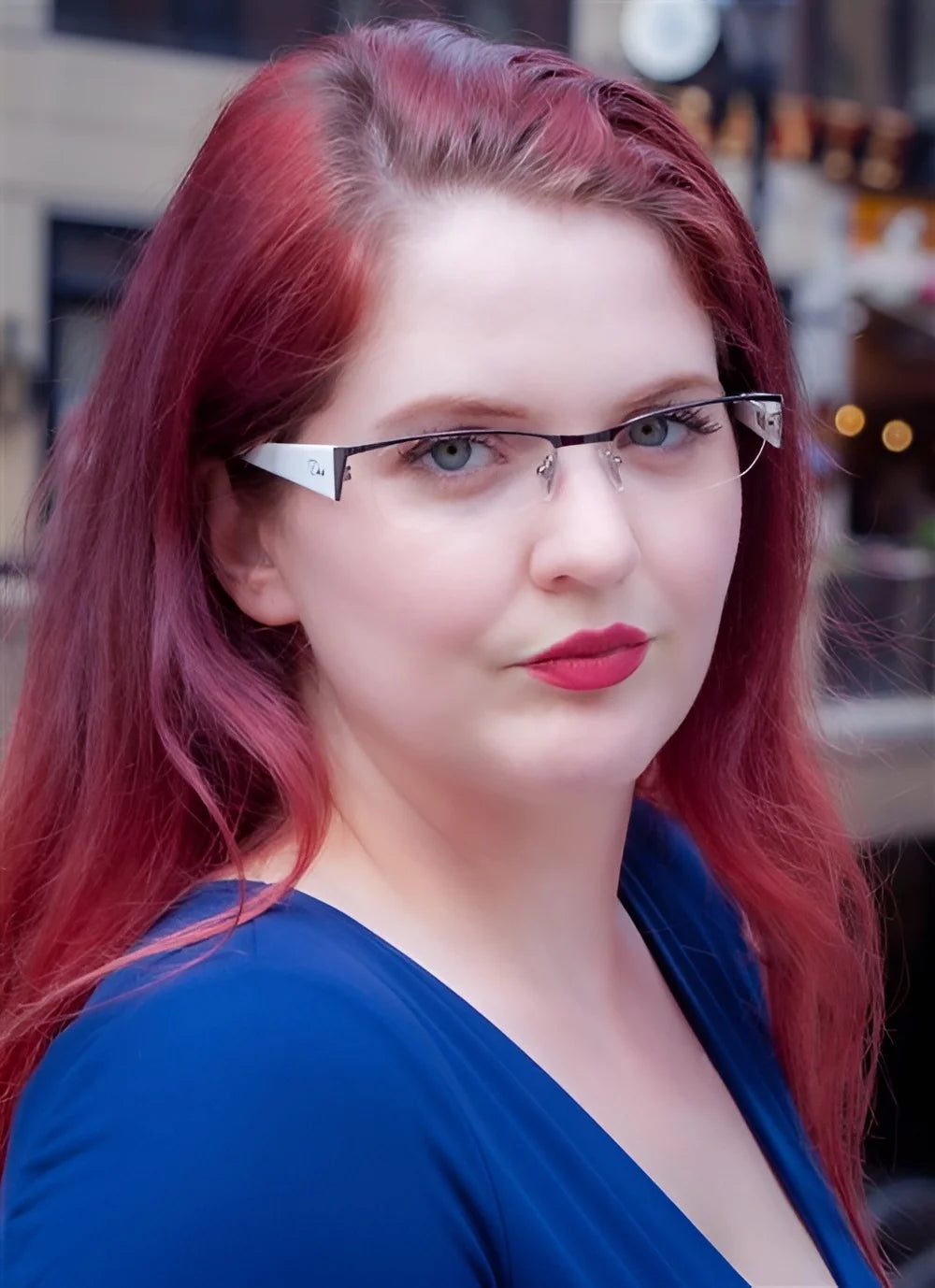 A person with long red hair and DEA Eyewear BIBIANA eyeglasses in size 54, dressed in a blue top, stands outdoors.