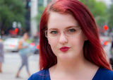 A woman with vibrant red hair and chic DEA Eyewear CATALINA eyeglasses is standing outdoors, the busy city street bustling in the background.