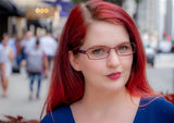 A stylish woman with red hair and glasses stands on a city street, wearing a blue top that perfectly complements her DEA Eyewear LILLY Eyeglasses in Size 52.