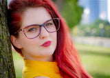 A person with red hair and DEA Preferred BARI eyeglasses, size 60, leans against a tree in the park, wearing a yellow top and looking directly at the camera.