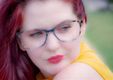 A person with red hair wearing DEA Preferred BARI Eyeglasses (Size 60) and a yellow top is wistfully gazing to the side in a soft-focus outdoor setting.