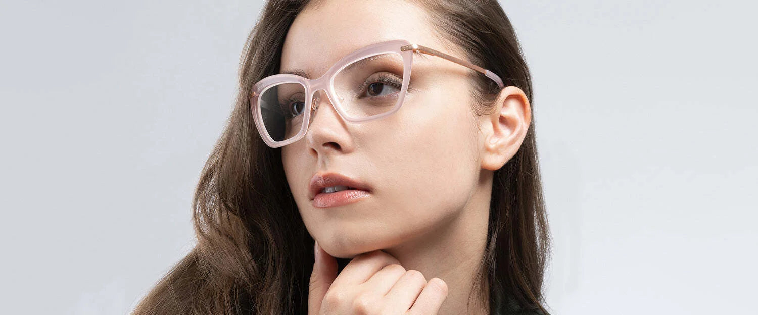 A woman with long brown hair wears stylish pink Gunnar Anna Computer Glasses, Size 54. The cat-eye frames have a hint of curiosity in her gaze, featuring advanced GUNNAR lens technology for blue light blocking as she thoughtfully touches her chin.