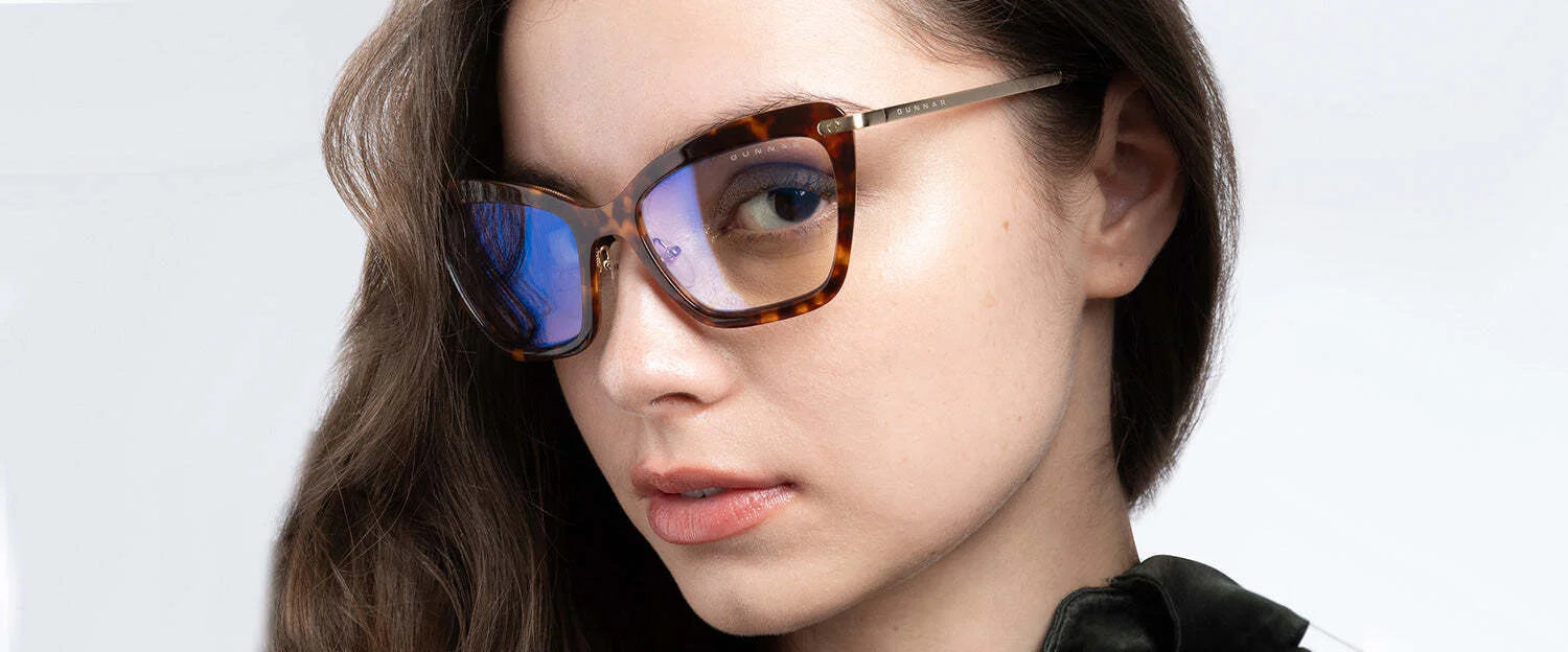 Wearing Gunnar Anna Computer Glasses (Size 54), a woman confidently gazes at the camera, flaunting stylish cat-eye frames and tortoiseshell glasses with blue lenses, featuring cutting-edge GUNNAR lens technology.