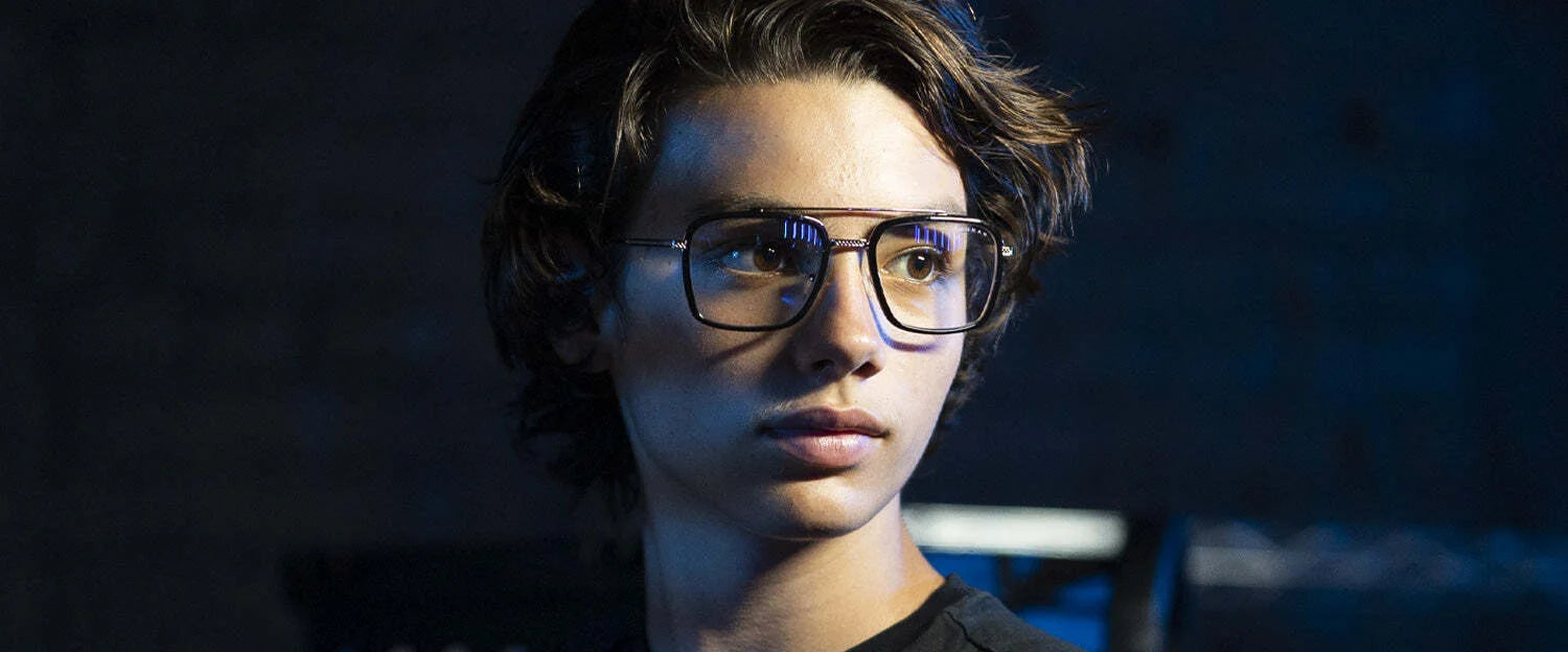 In a dimly lit setting, a person with wavy hair glances sideways, wearing Gunnar Barron Computer Glasses featuring a sleek stainless steel frame.
