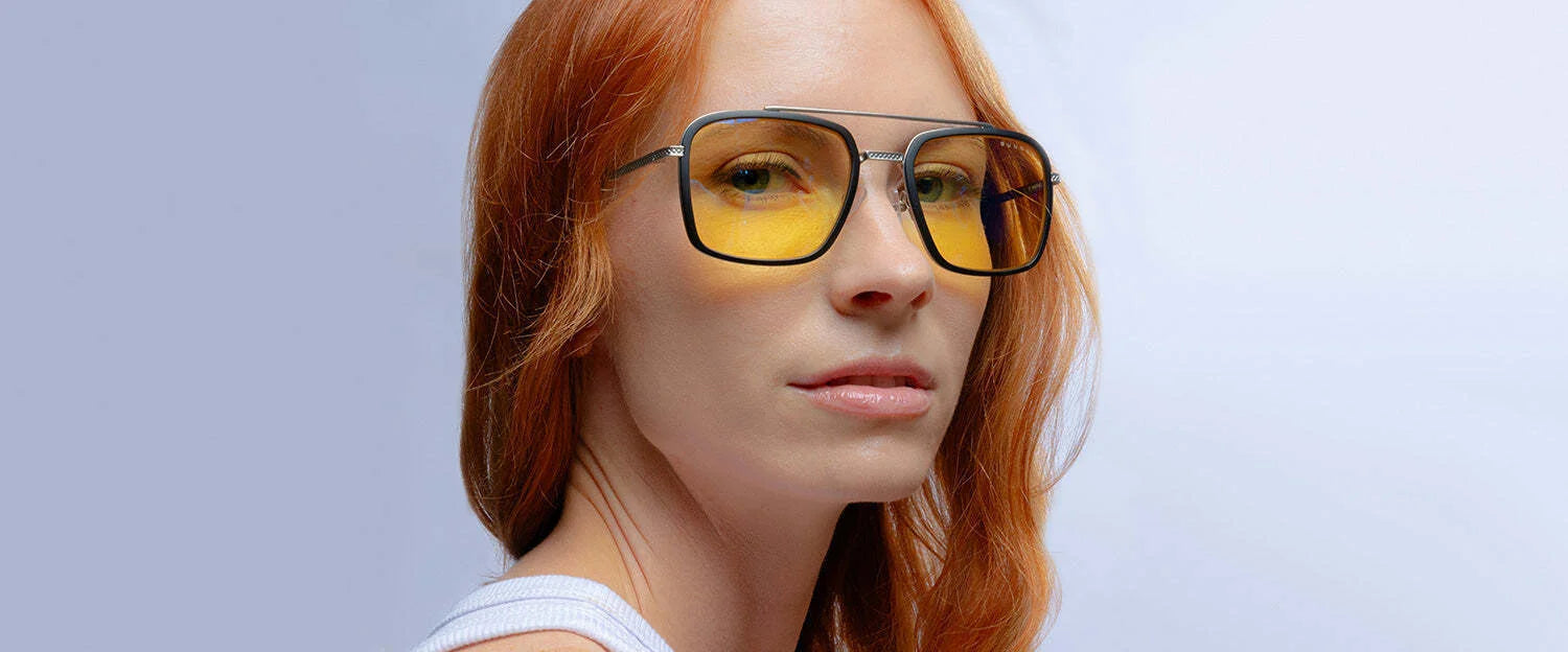 A red-haired individual wears Gunnar Barron Computer Glasses Size 55, featuring yellow lenses and a sleek stainless steel frame, set against a light blue background.