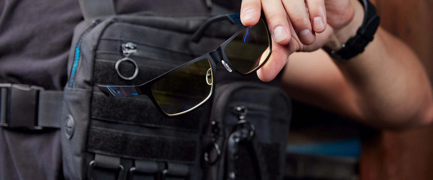 In an outdoor setting, a person in a tactical vest confidently holds Gunnar Call of Duty Covert Edition Computer Glasses. These black sunglasses feature the latest Gunnar patented lens technology, perfect for any mission.