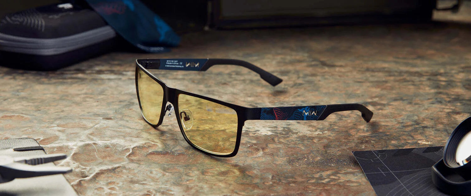 Gunnar Call of Duty Covert Edition Computer Glasses, with yellow-tinted rectangular lenses and black frames, rest on a textured surface near various accessories. They feature Gunnar's patented lens technology for effective blue light blocking.