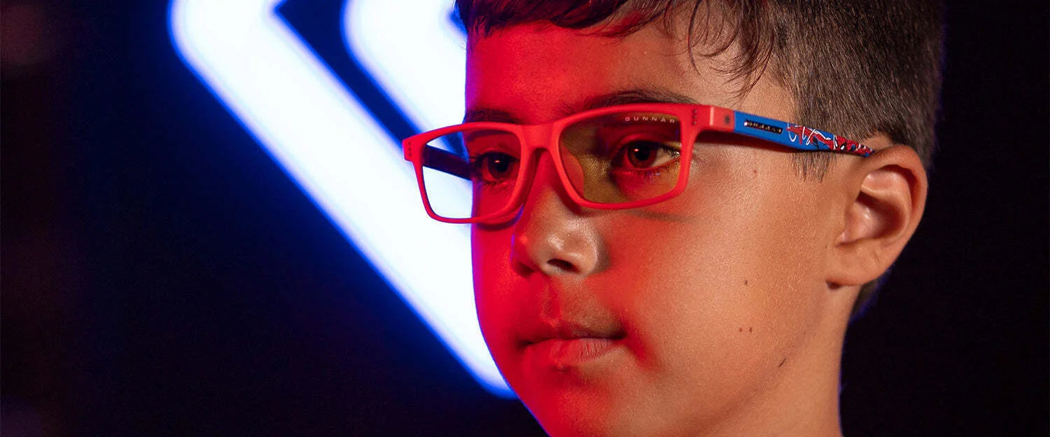 A child wears Gunnar Cruz Kids Spider-Man Edition Computer Glasses, featuring vibrant frames bathed in red with a striking blue neon shape behind them.