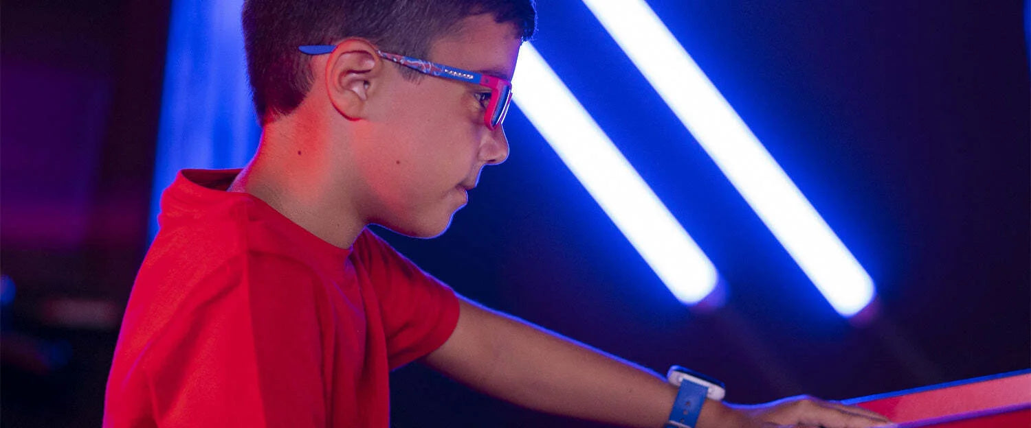 A boy in a red shirt and Gunnar Cruz Kids Spider-Man Edition Computer Glasses is deeply engrossed in a task, with blue neon lights casting a glow behind him.